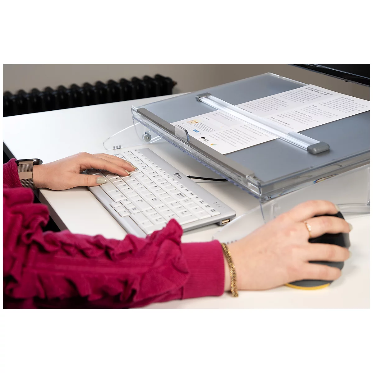 Person using keyboard and mouse at a desk. A document lies on a lectern. Arm with a golden bracelet.