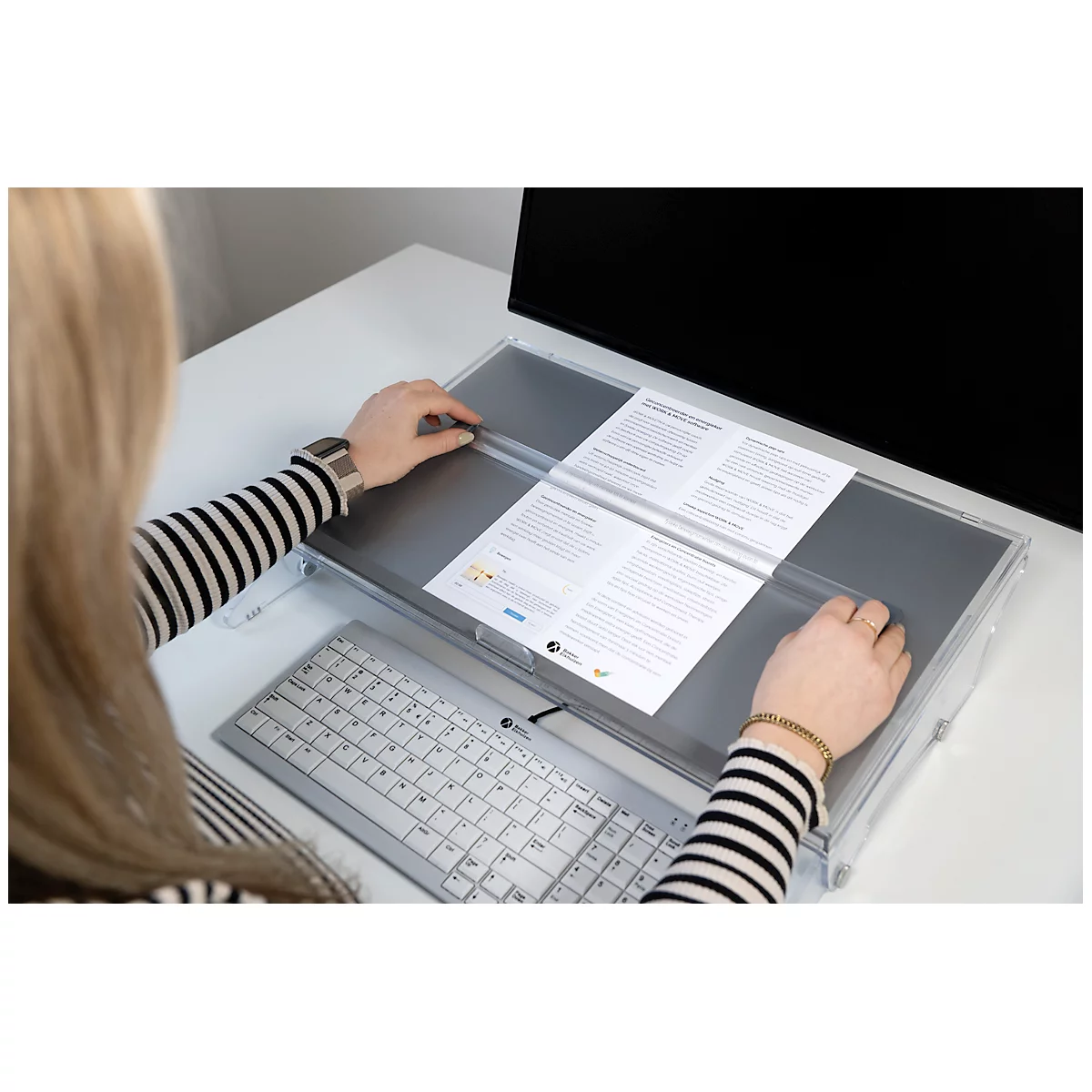Woman at desk, reading documents placed in a clear holder in front of the monitor.