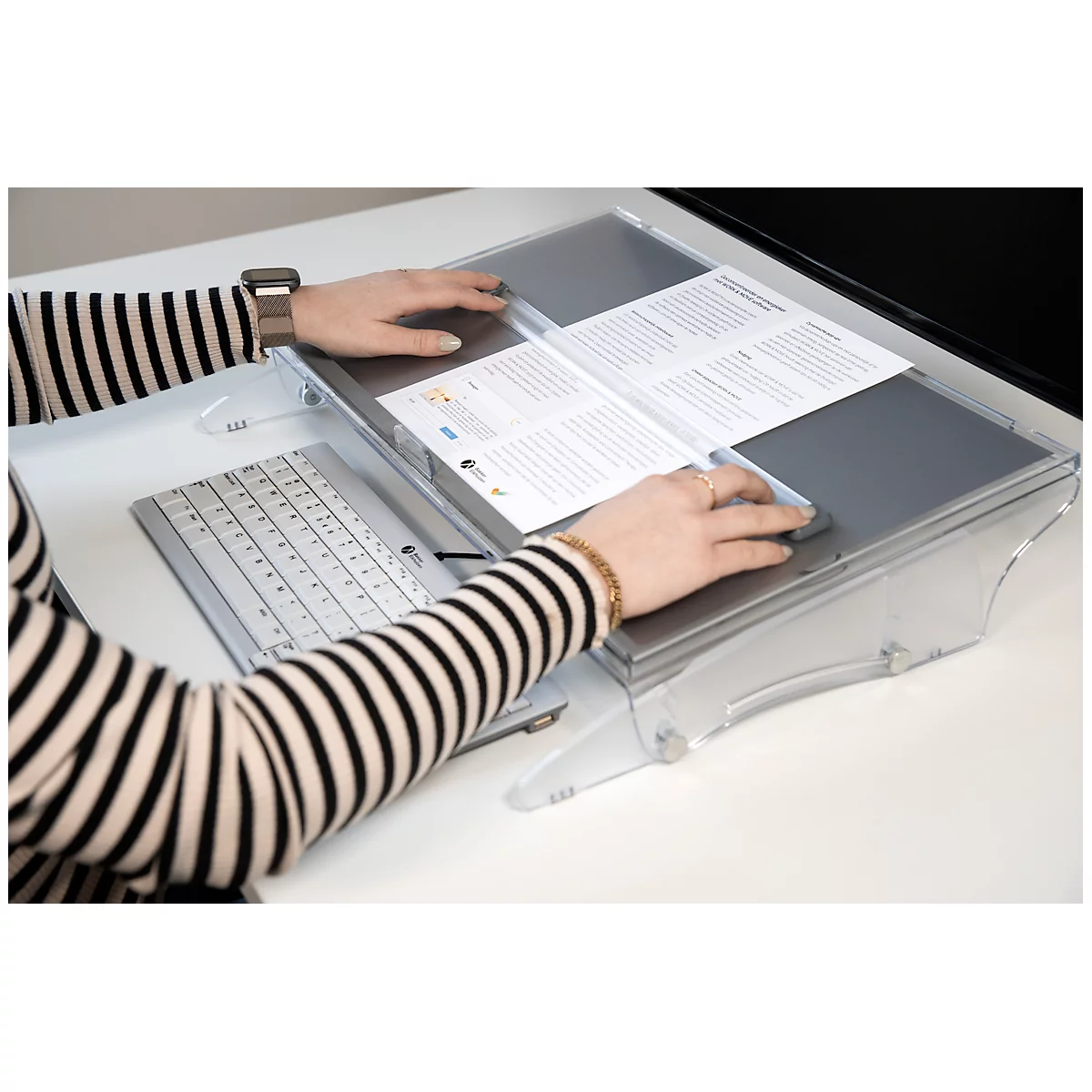 Person wearing a ring, wristwatch, and bracelet operates a special reading device with a keyboard and mouse. There is paper on the reading device.