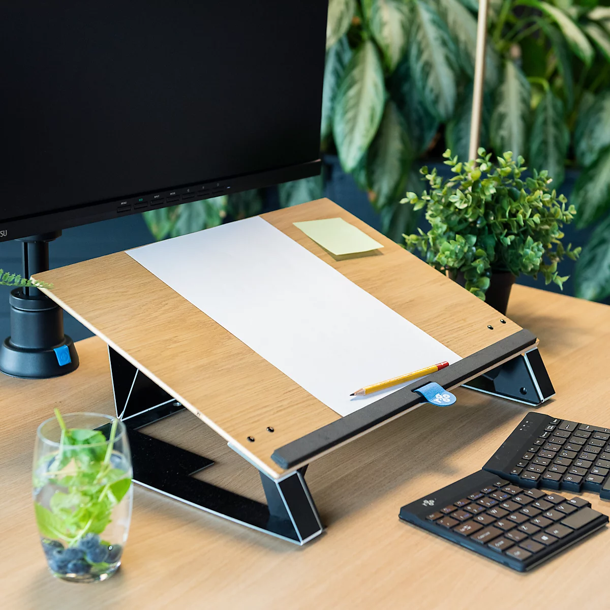 Wooden laptop stand with paper and pencil. Next to it a keyboard and a glass of water with blueberries and mint.