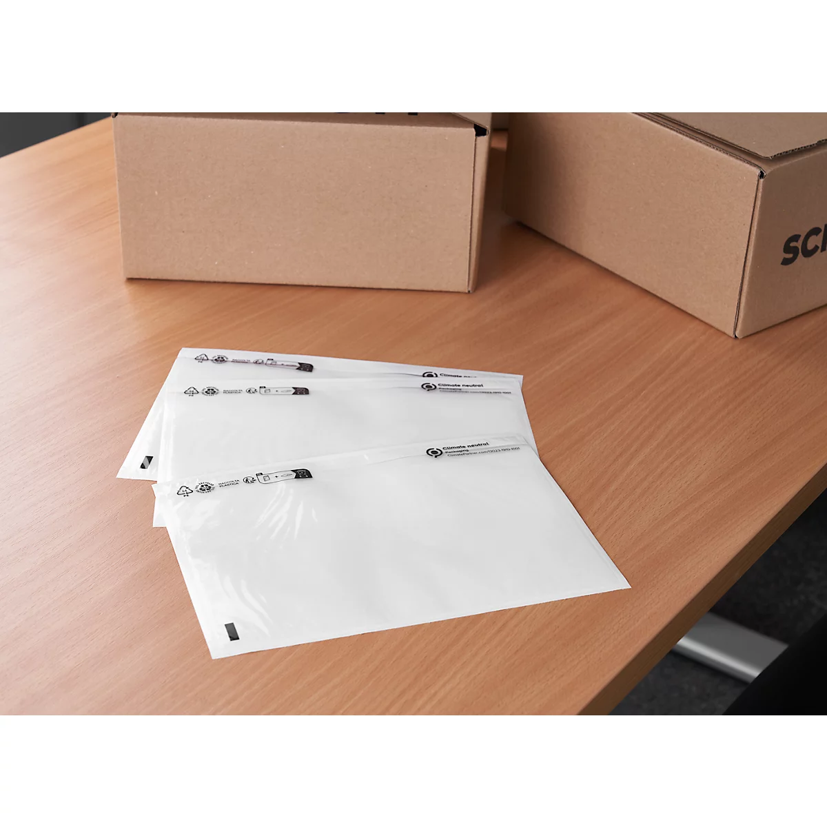 Three transparent envelopes lie on a wooden table. Cardboard boxes in the background.