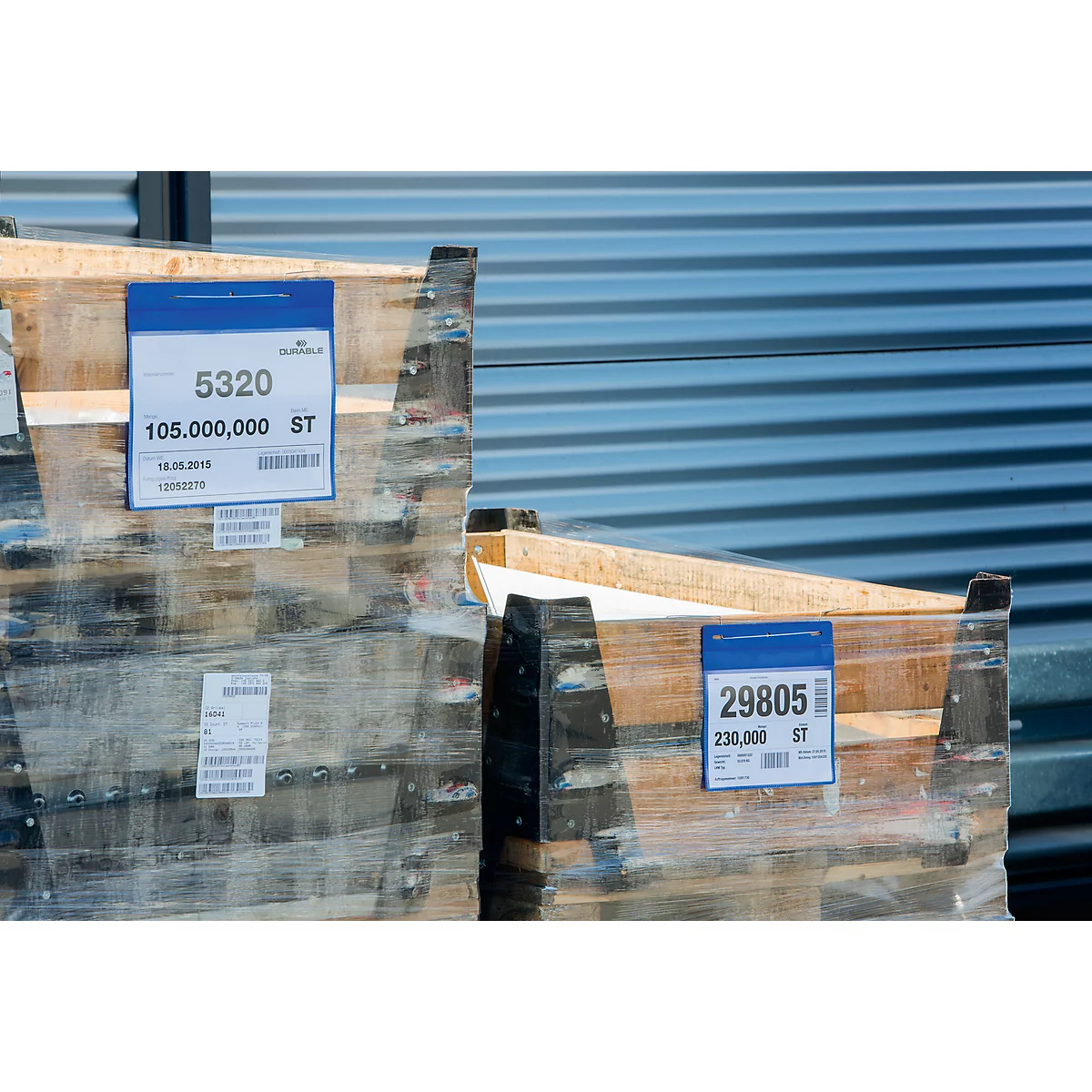 Zwei Paletten mit Holzaufbauten, eingeschweißt in Folie. Blaue Etiketten mit Ziffern und Barcodes sind angebracht.