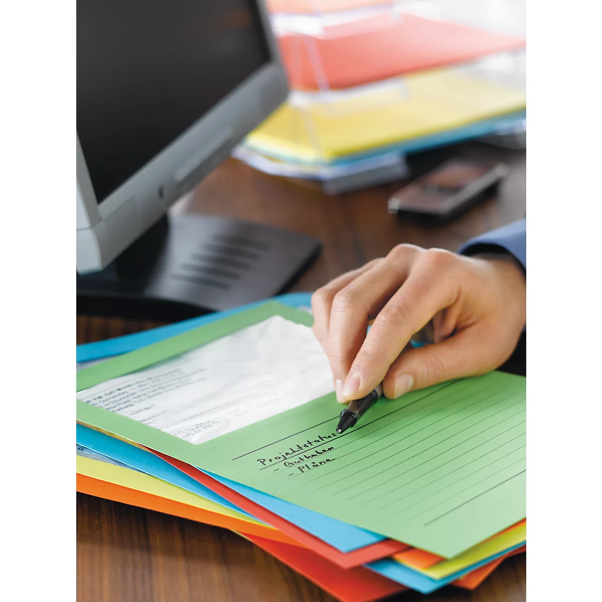Une personne écrit sur un dossier vert. En dessous se trouvent d'autres piles de dossiers colorés, un ordinateur et un téléphone portable en arrière-plan.