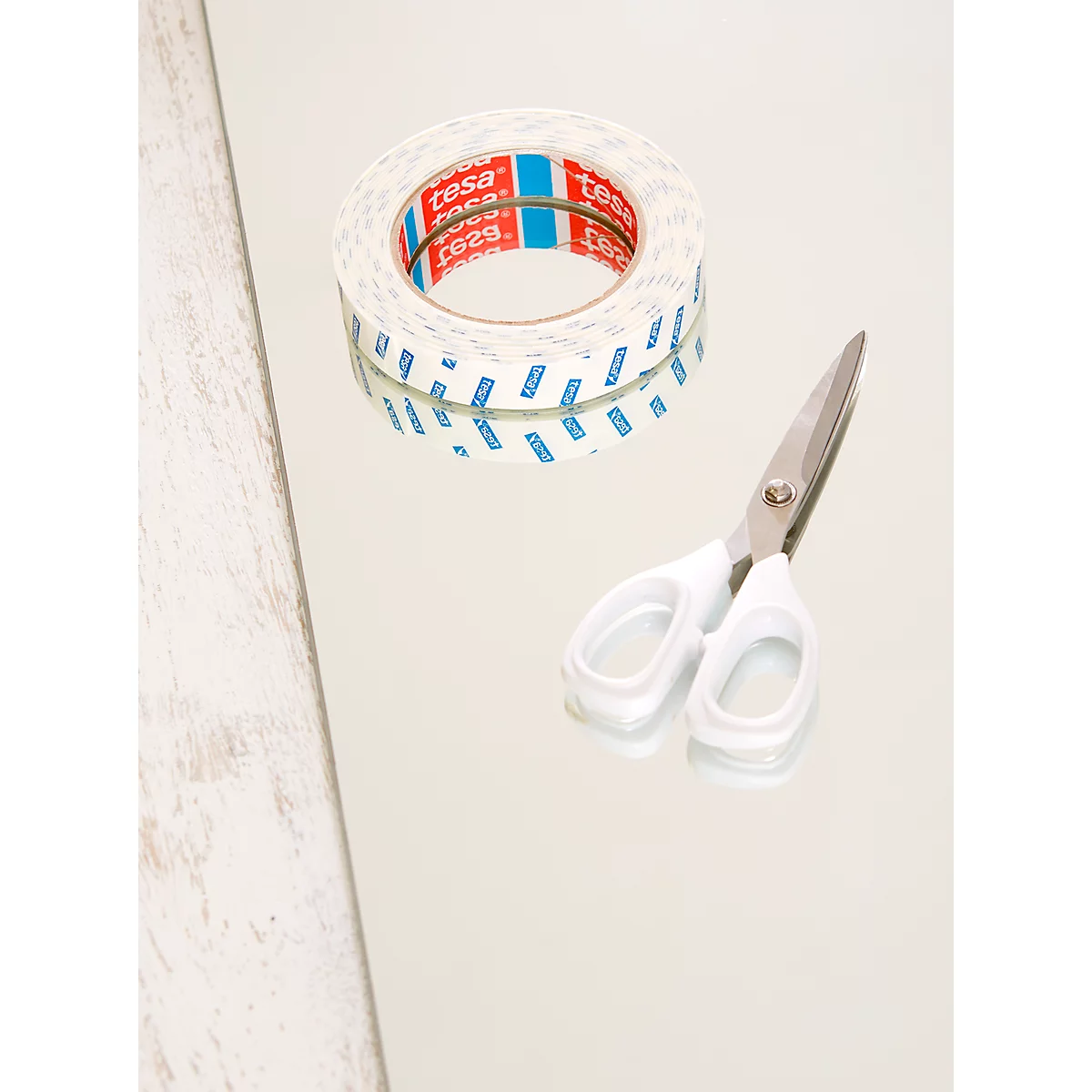 A roll of double-sided tape with red and blue markings and white scissors lie on a smooth surface.