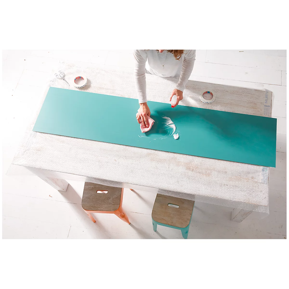 Person wipes on a turquoise surface with a sponge. Underneath a wooden table, next to it two stools.