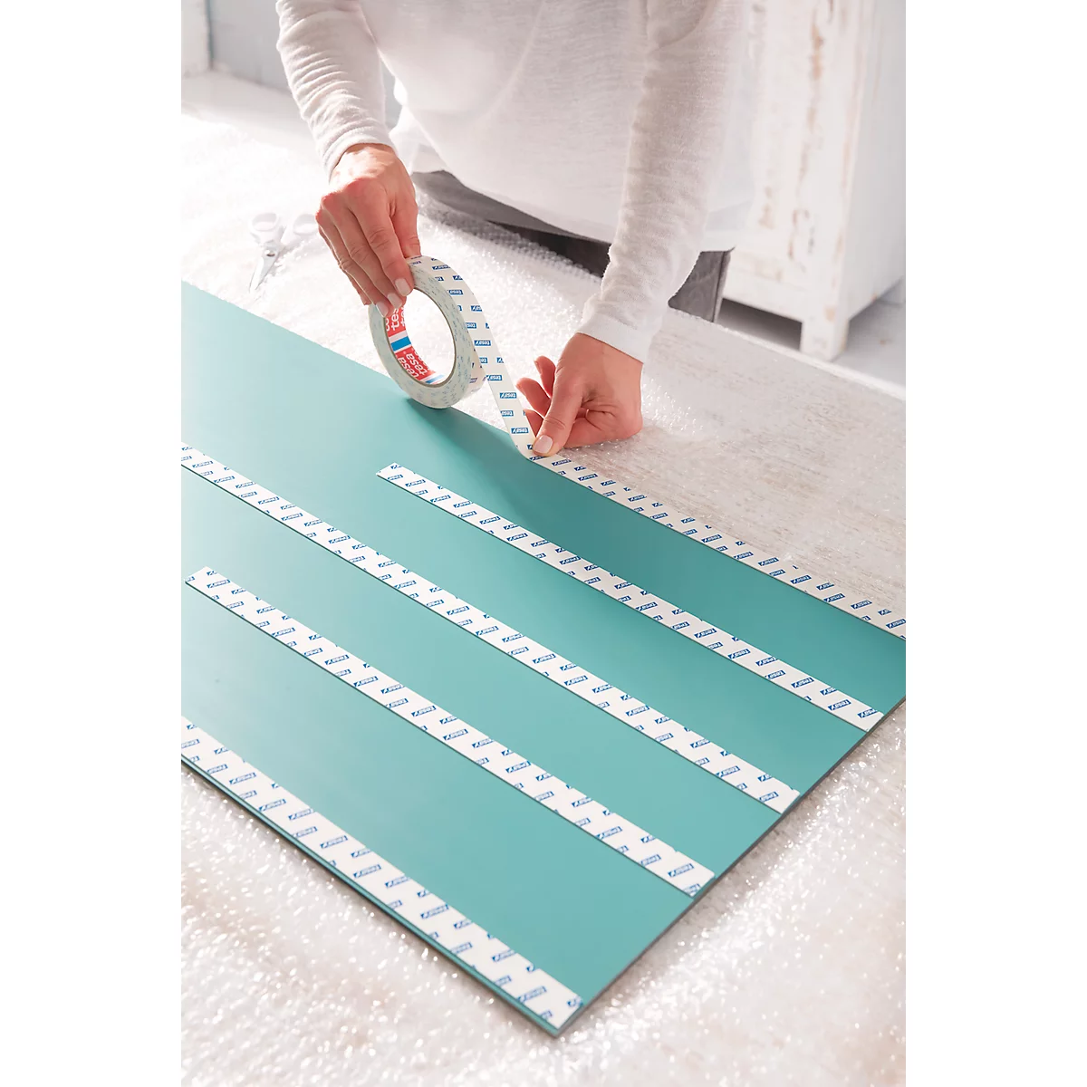 Hands are applying double-sided tape to a turquoise board.