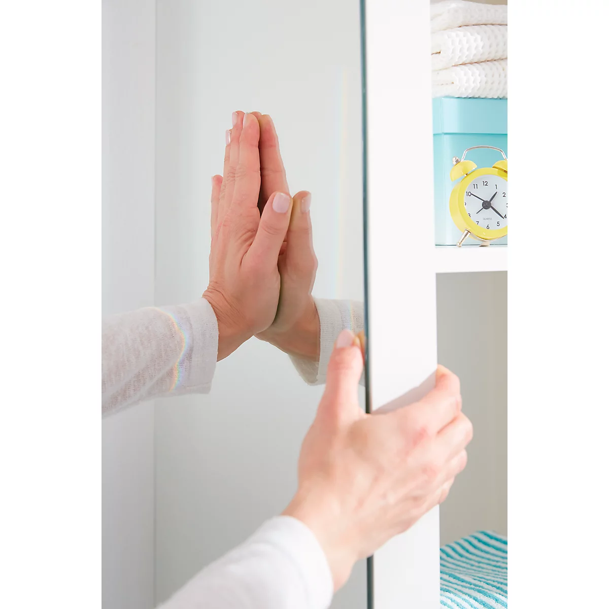 Hands touching a mirrored door located on a white cabinet. In the background there is a shelf with towels, a box and a clock.