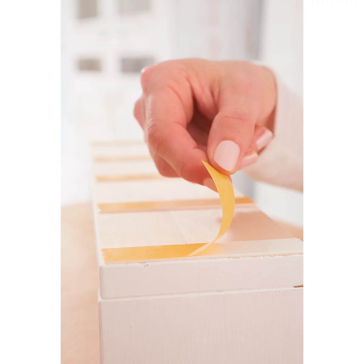 A hand is removing yellow tape from a white wooden box. Light brown tapes are attached to the box.
