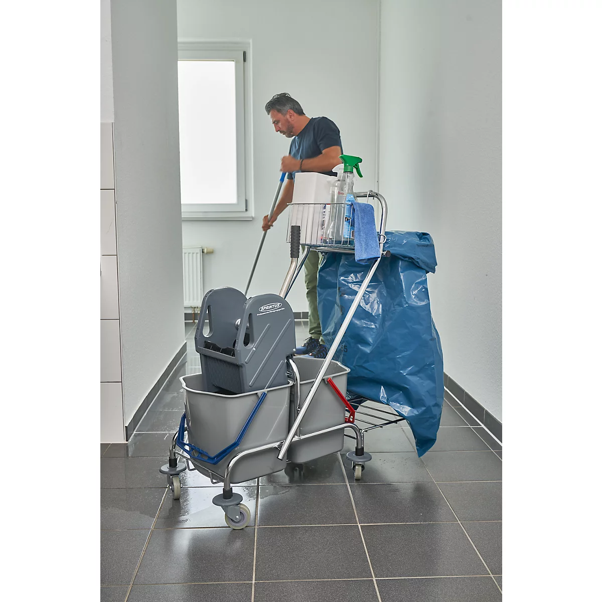 Man cleaning the floor in a corridor with a cleaning cart. He stands in the middle of the hallway and uses a mop.