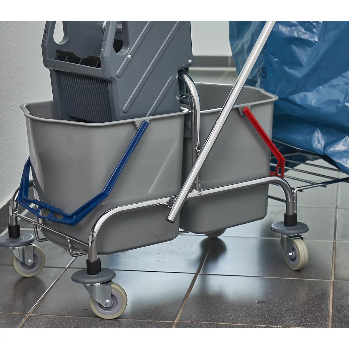 Grey cleaning cart with two buckets, blue and red handles. Behind the cart a blue garbage bag.