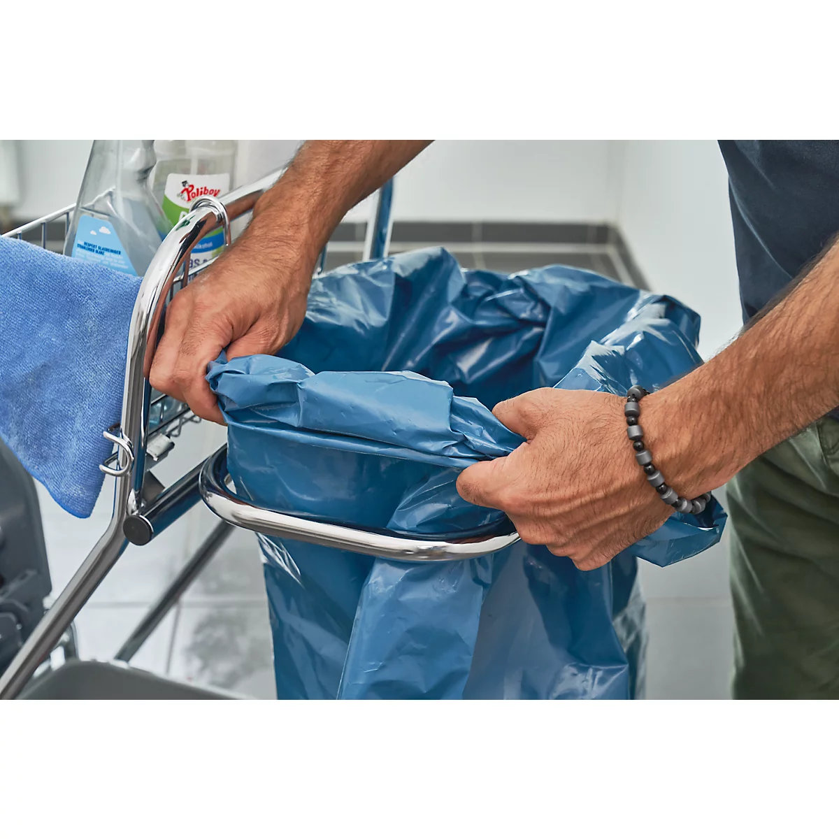 Man fixing a blue trash bag in a trash can. In the background a cart with towel and cleaning products.