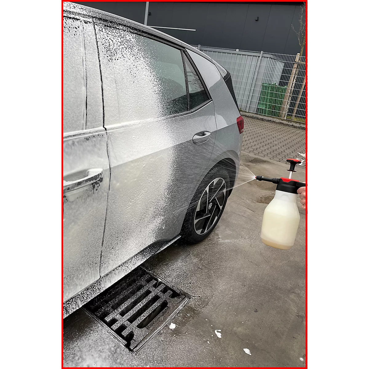 Ein graues Auto wird mit Schaum eingesprüht. Eine Hand hält eine Sprühflasche. Auf dem Boden ist ein Gullydeckel zu sehen.