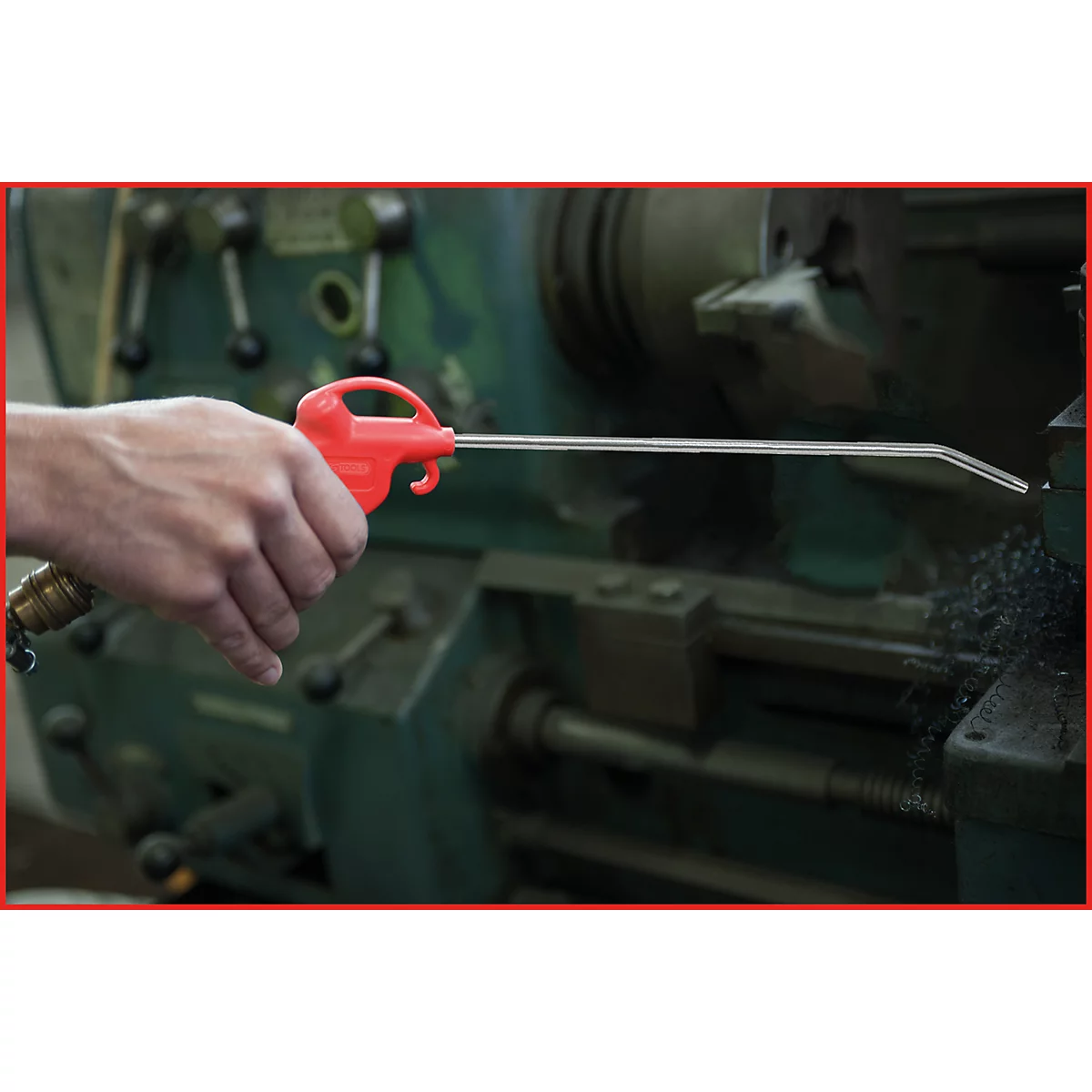 Eine Hand hält eine rote Druckluftpistole, die auf eine Maschine gerichtet ist. Metallspäne liegen daneben.