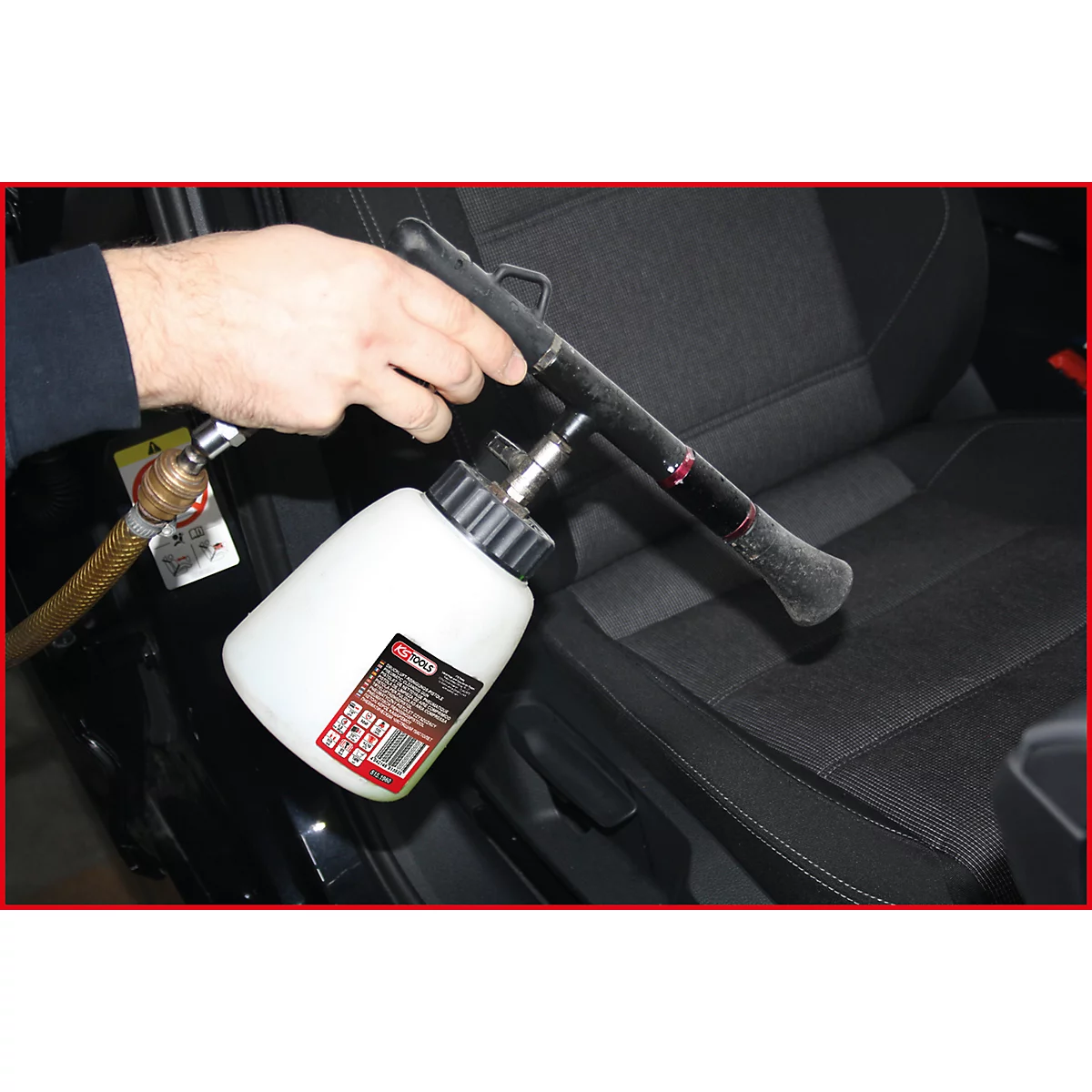Reinigungswerkzeug reinigt Autositze. Schwarzer Sitz, weiße Flasche, schwarzes Werkzeug. Hand hält Werkzeug, Schlauch angeschlossen.