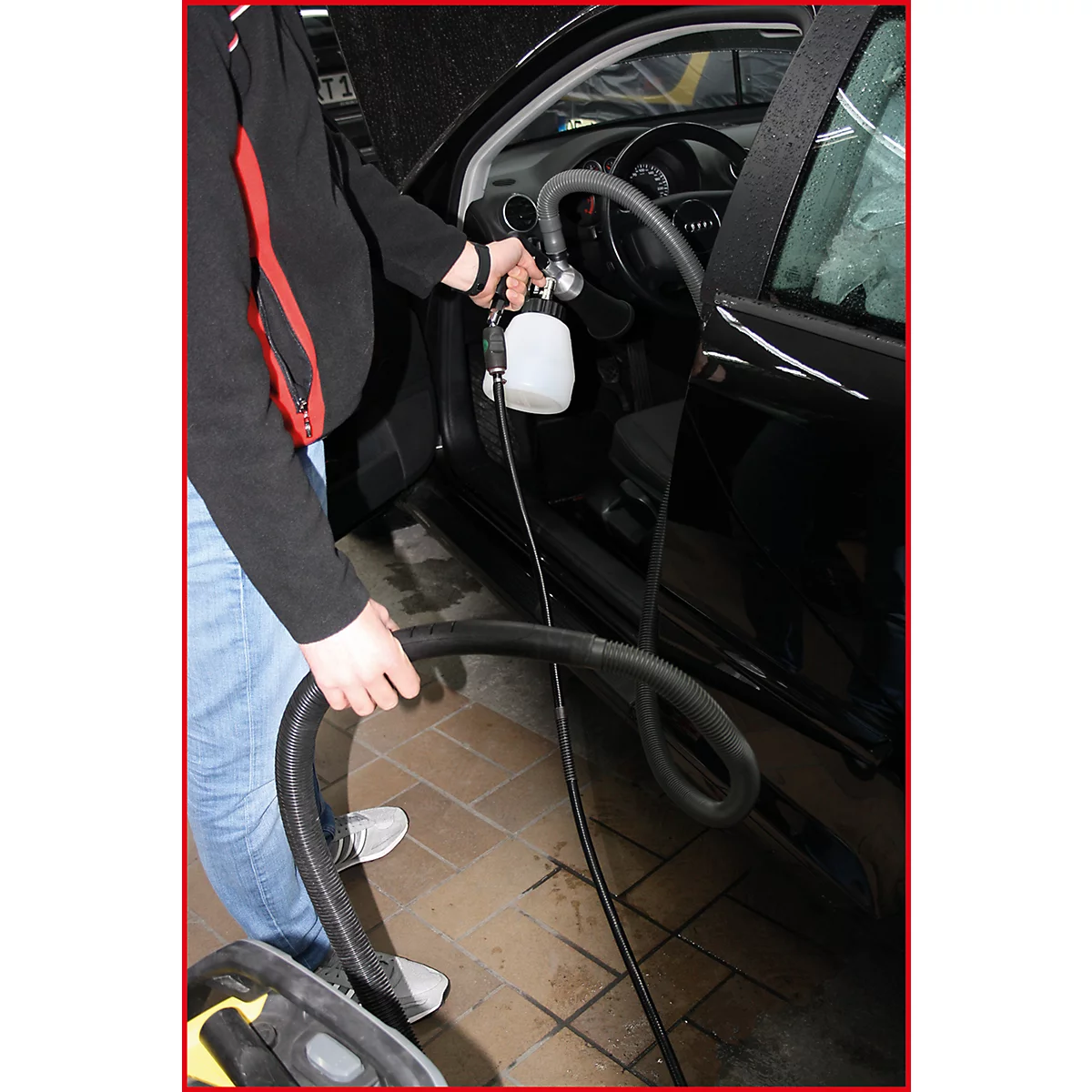 Ein Mann reinigt den Innenraum eines schwarzen Autos. Er hält einen Schlauch und eine Sprühflasche.