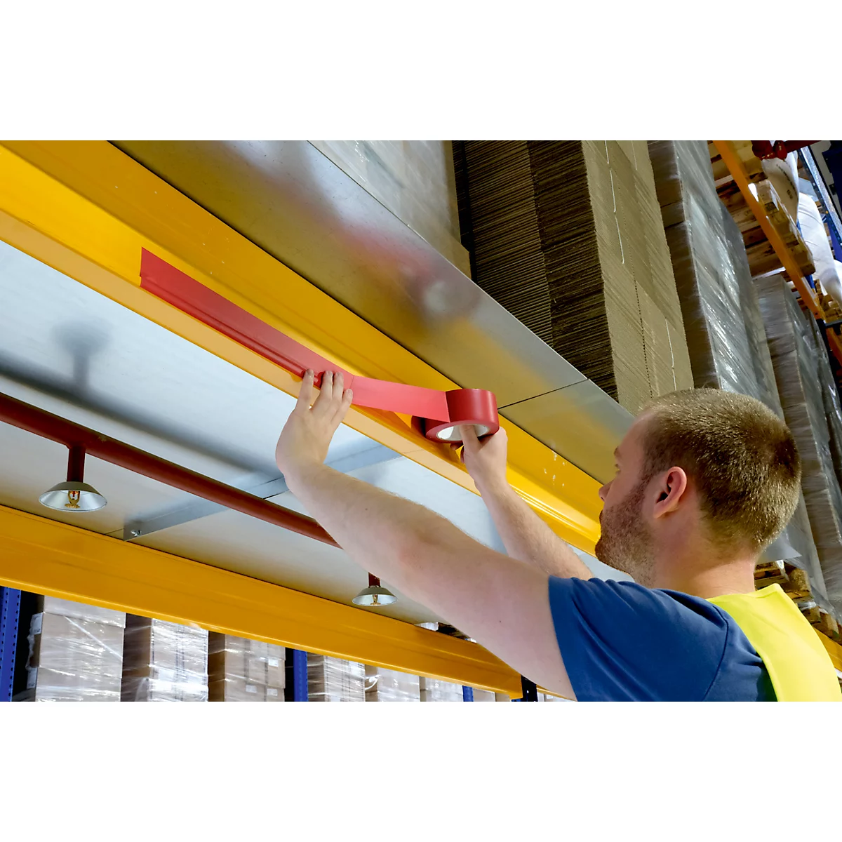 A man attaches red tape to a yellow metal beam. Shelves with goods can be seen in the background.