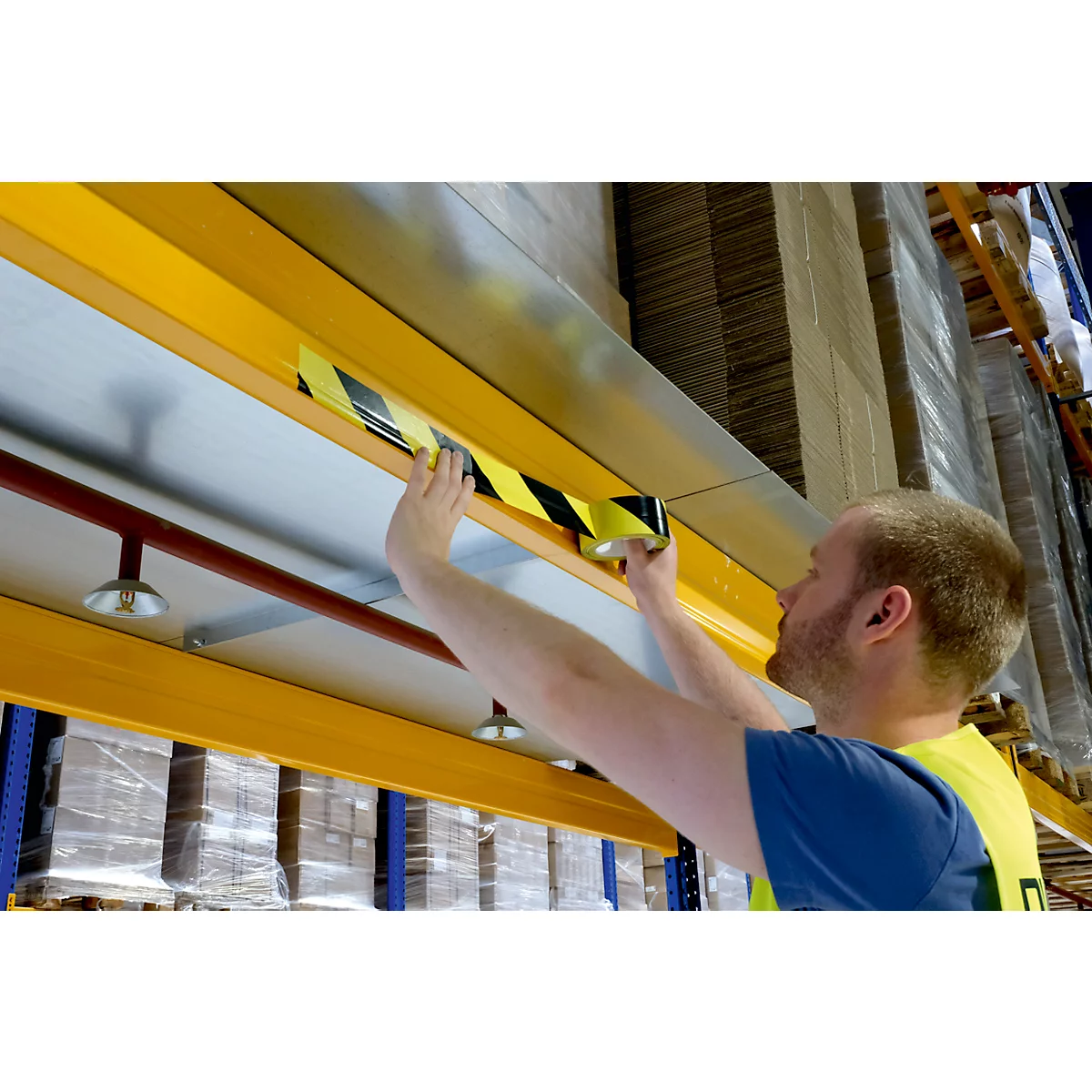 A man is applying a black and yellow warning tape to a yellow beam in a warehouse.