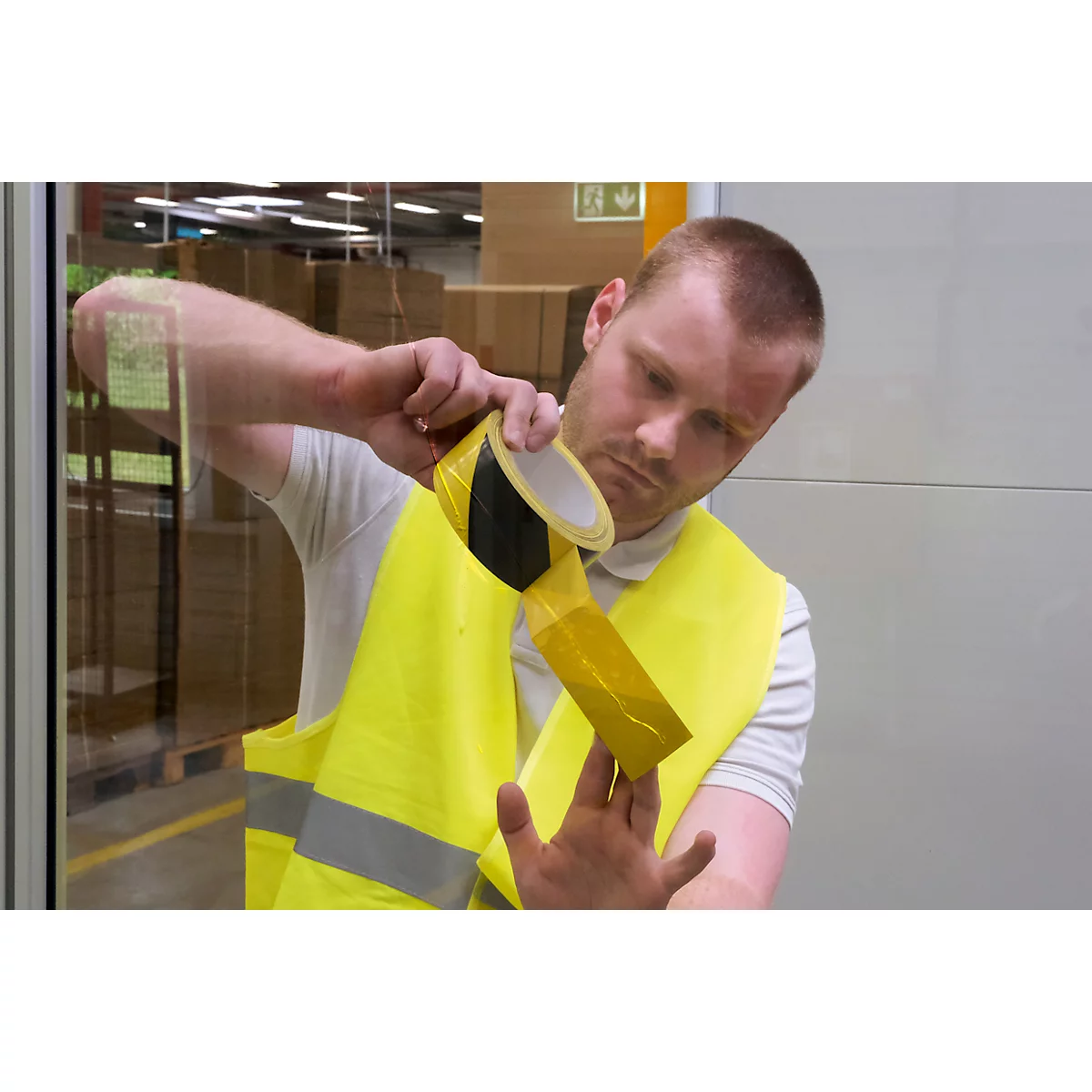 A man is applying yellow warning tape to a yellow safety vest. He is holding the roll and the vest. In the background a warehouse.