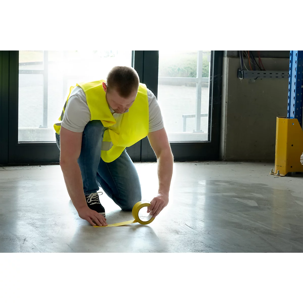 A man kneels and applies yellow tape to a concrete floor.