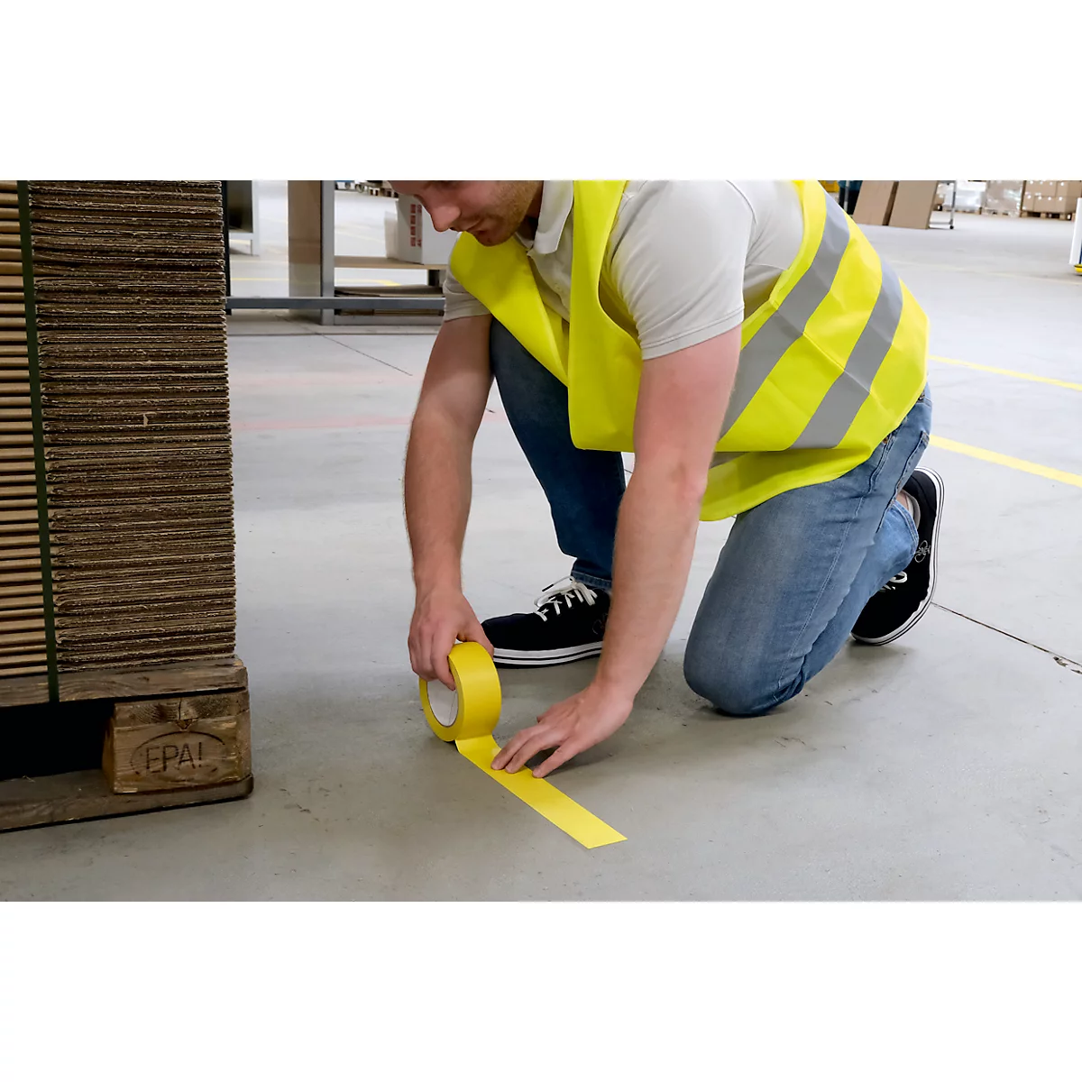 A man kneels and applies yellow tape to the concrete floor. He wears a yellow safety vest.