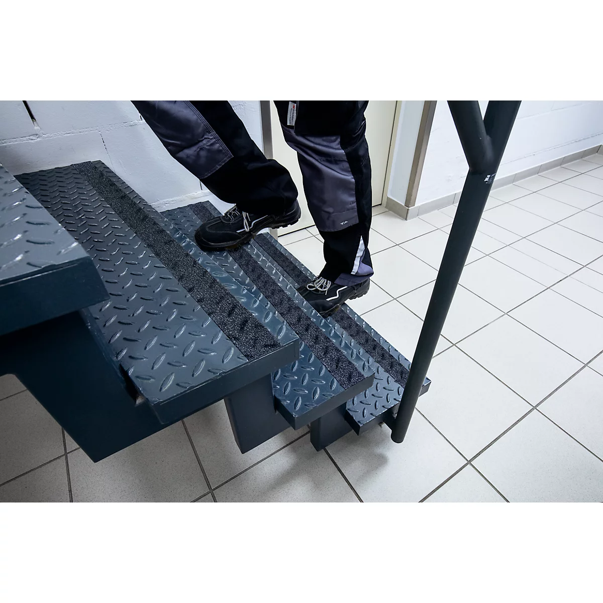 Person climbing a metal staircase. Grey steps with diamond pattern. Trousers, shoes, black handrail.