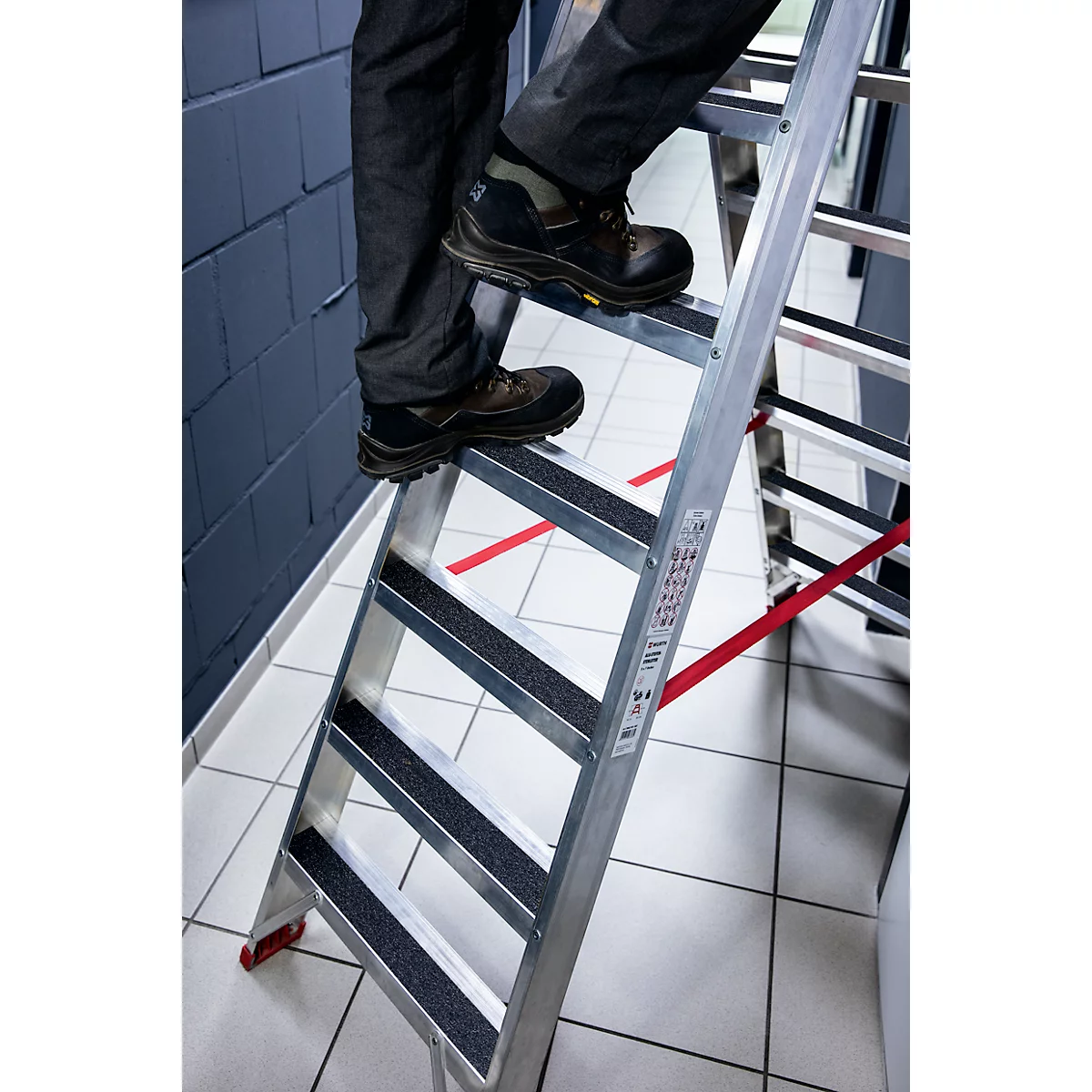Person is climbing a silver ladder. Black shoes on non-slip steps. Red stripe on the floor.