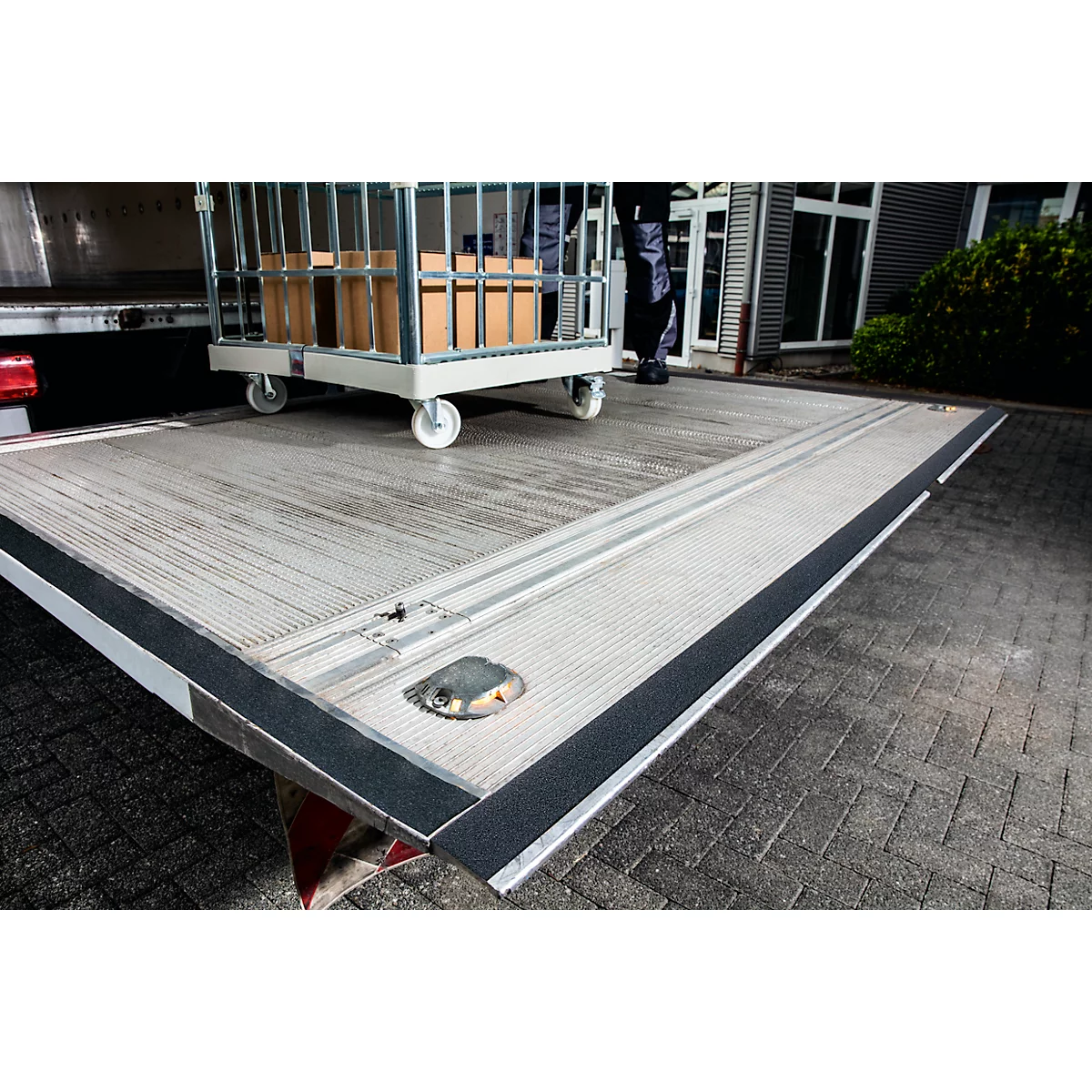 Loading ramp of a truck with a loaded trolley. Black edge protection, paved floor, person in the background.