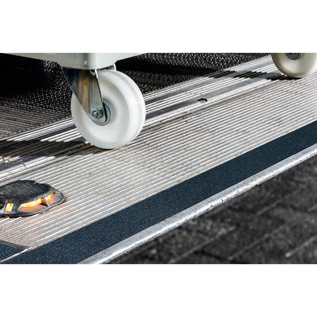 Close-up of a wheelchair ramp featuring a white wheel and a black non-slip strip. The surface is made of corrugated metal.