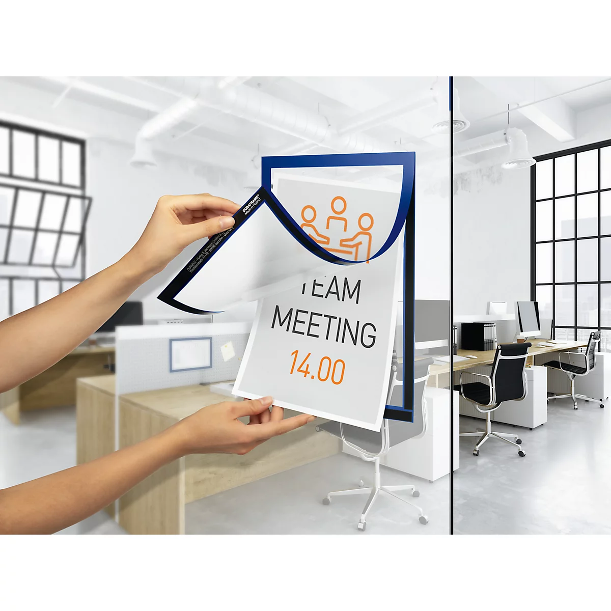 Person holding a sign with "Team Meeting 14.00" in a blue frame. An office in the background.
