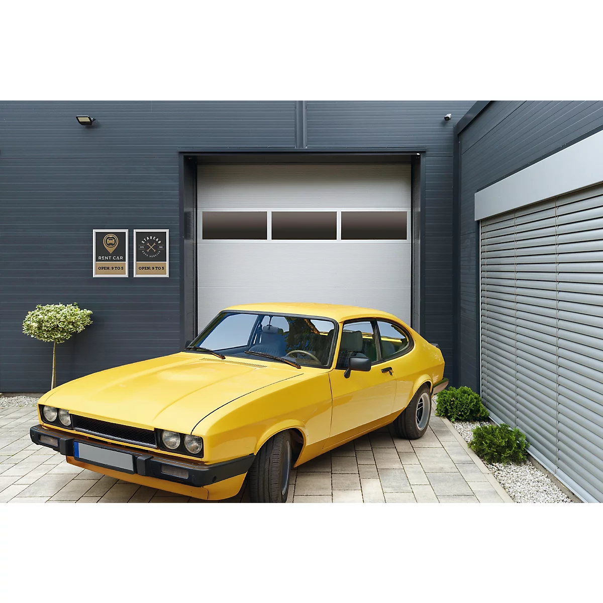 Ein gelbes Oldtimer-Coupé steht auf einer gepflasterten Einfahrt vor einem modernen Garagentor mit Lichtband.