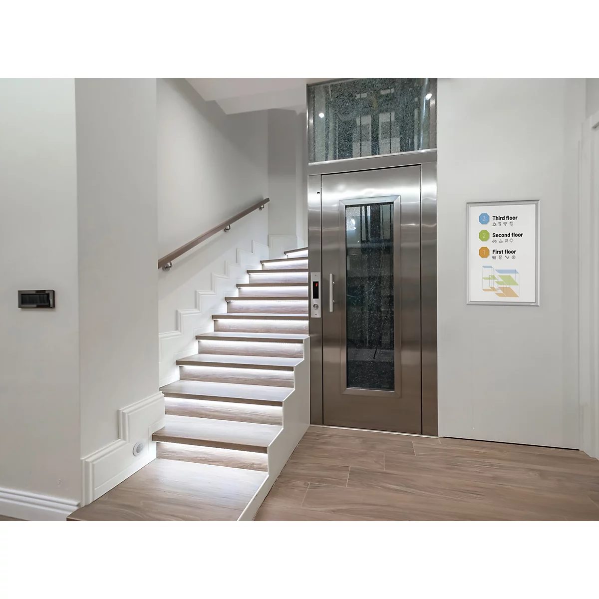 Moderne roestvrijstalen lift naast een verlichte houten trap in een licht wooninterieur.