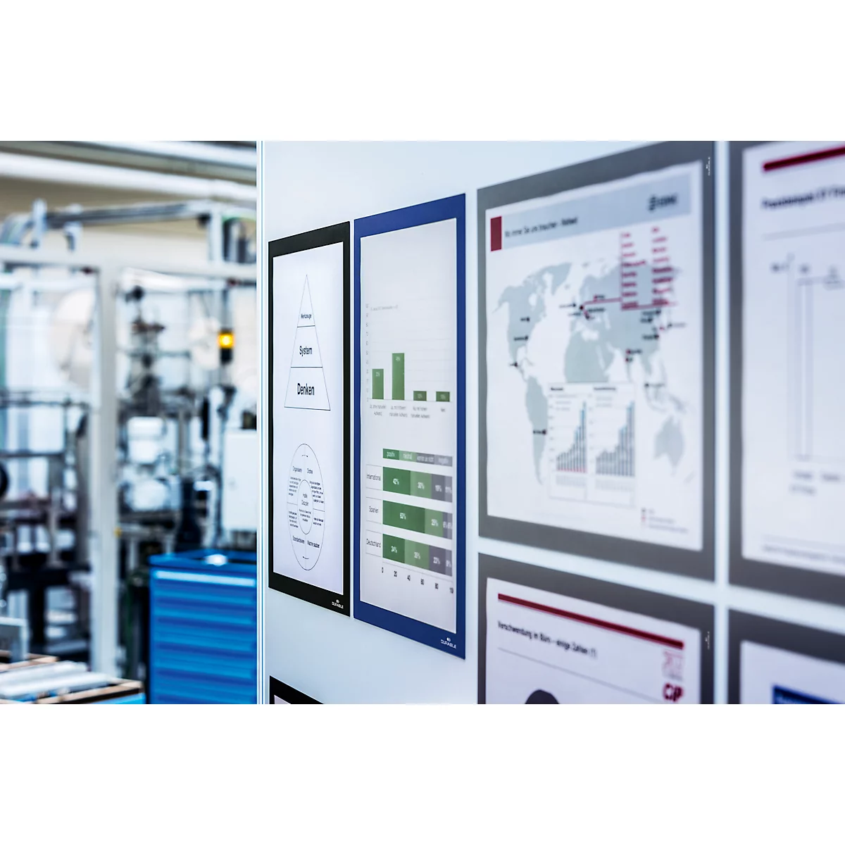Several framed diagrams and charts on a white wall. A factory in the background.
