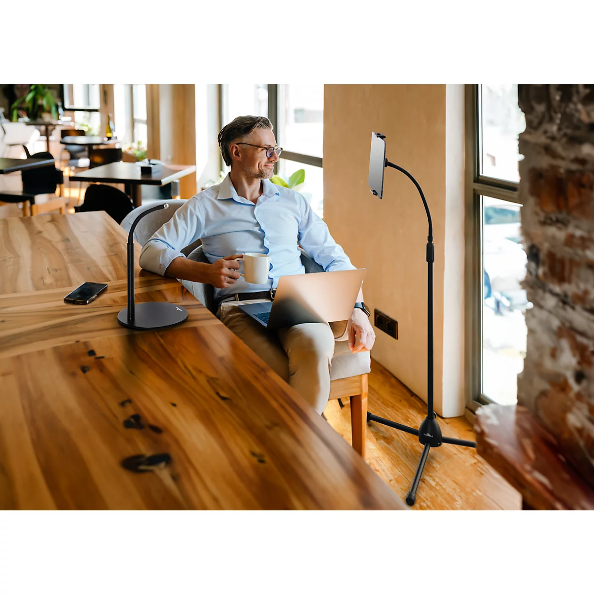 A man sits in an armchair with a laptop and a cup. A tablet is attached to a stand, the man is looking out of the window.