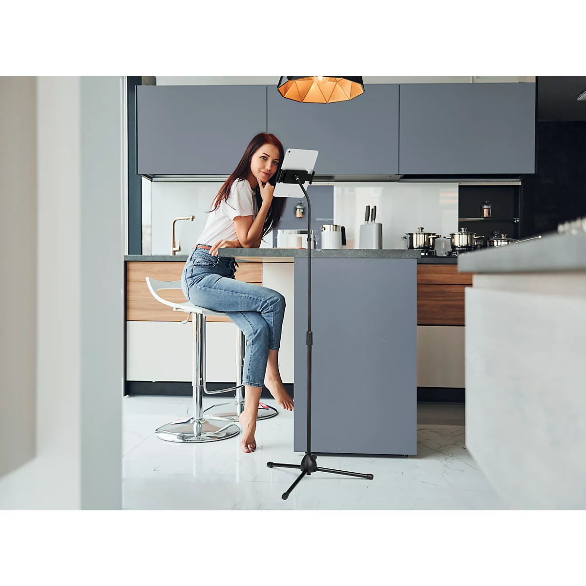 Young woman sitting at kitchen counter. She rests her chin on her hand and looks into the camera. Next to her, a tablet on a tripod.