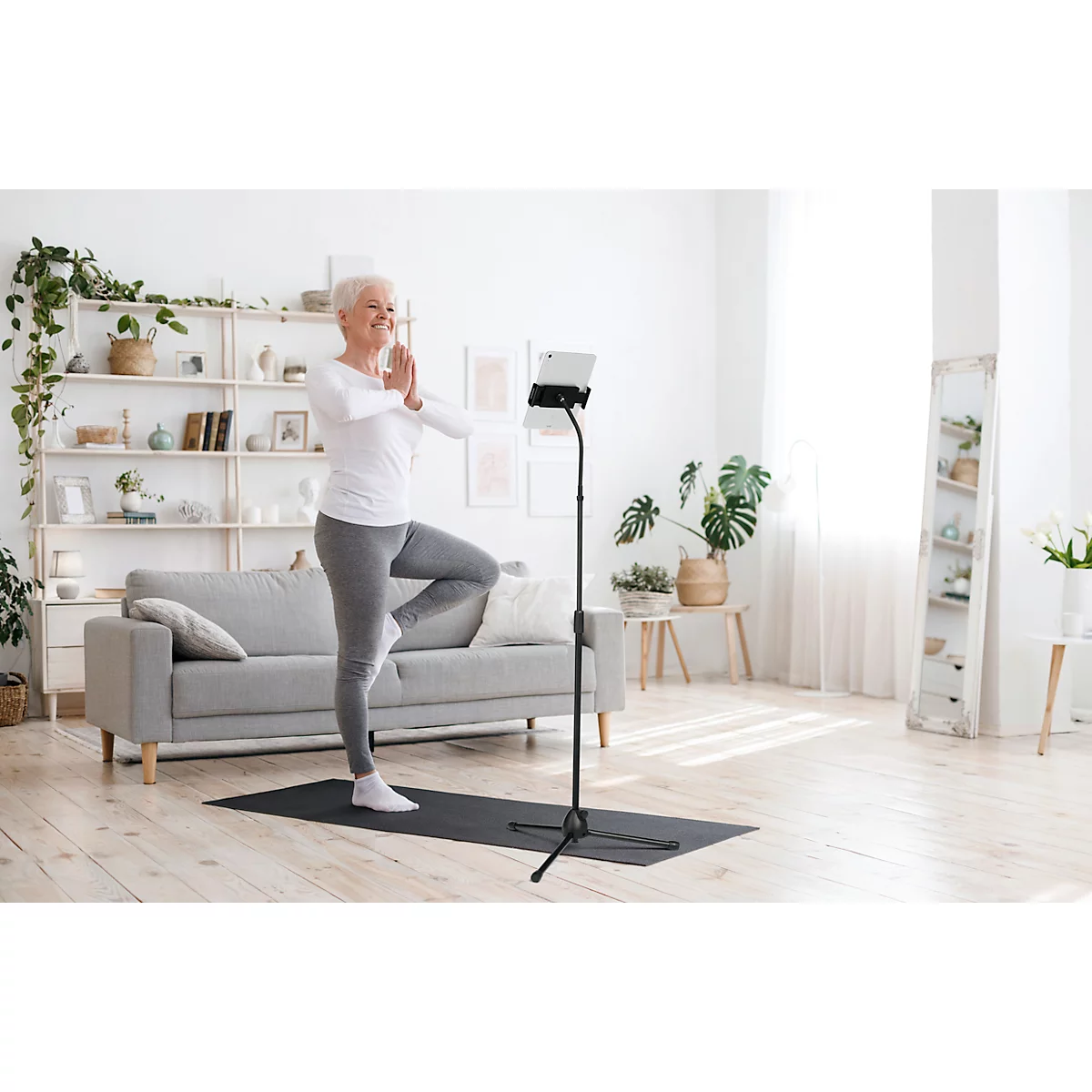 Senior woman doing yoga. She stands on one leg, arms in prayer position, in front of a tablet. Living room background.