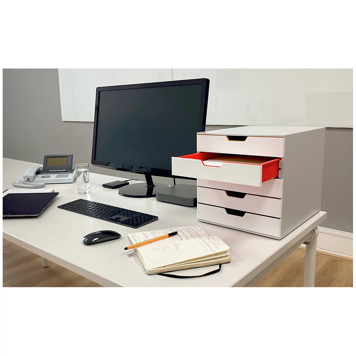 Desk with computer, drawer cabinet, phone, pen, and notebook. One drawer is open.