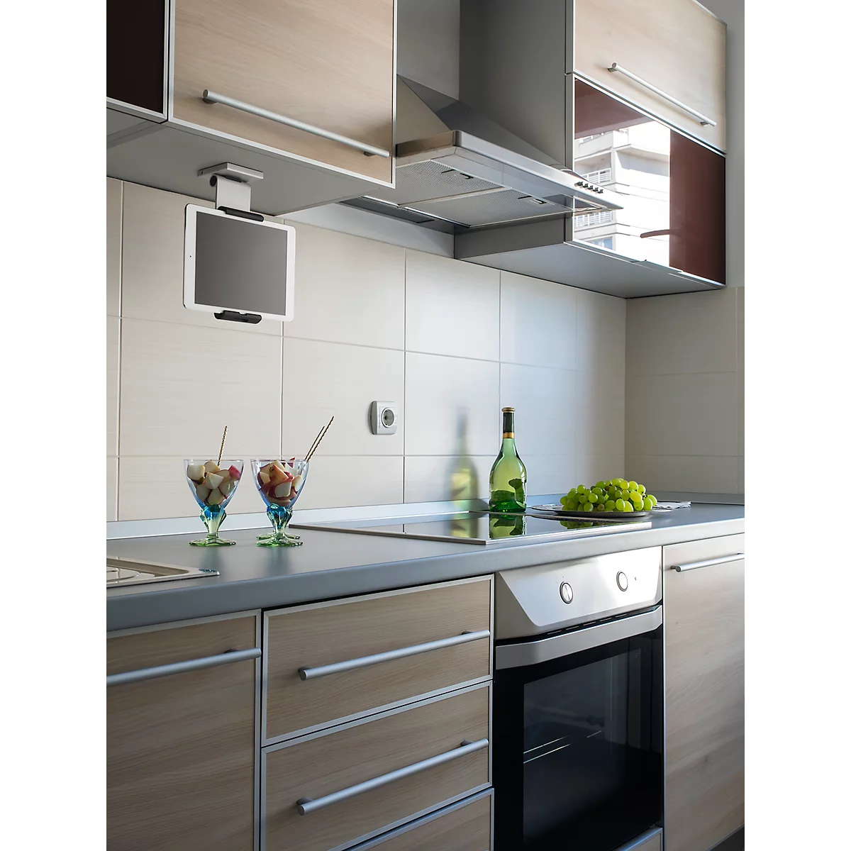 Modern kitchen featuring a tablet, fruit cups, and wine bottles. Light-colored cabinets, dark countertop, appliance buttons.