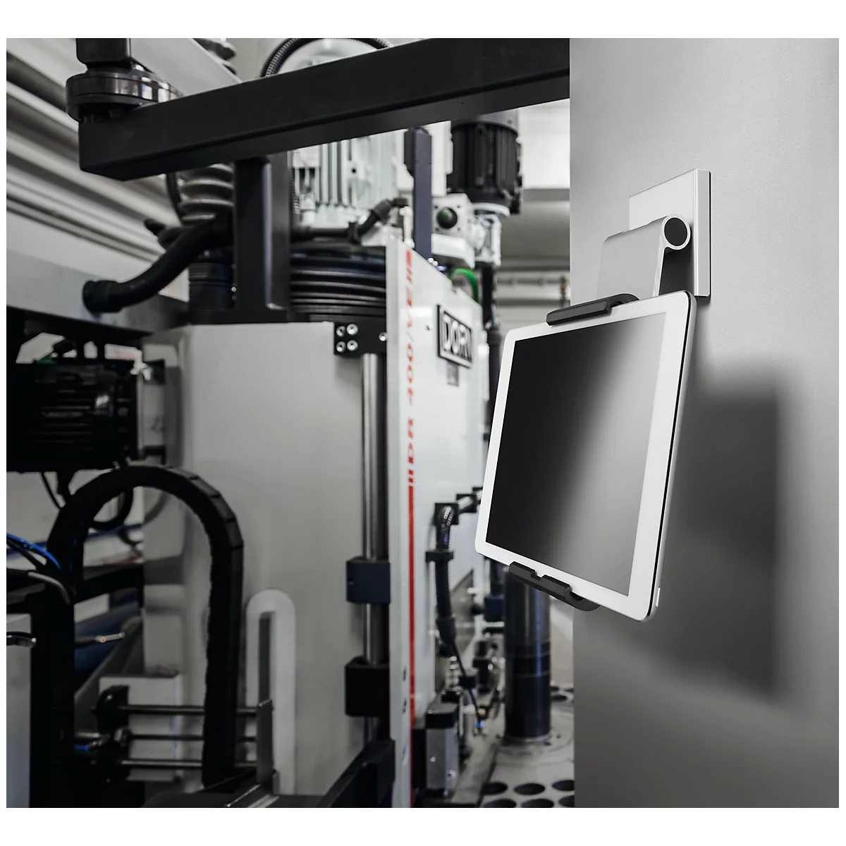Tablet on holder in front of industrial plant. Bright display, black holder, gray wall.
