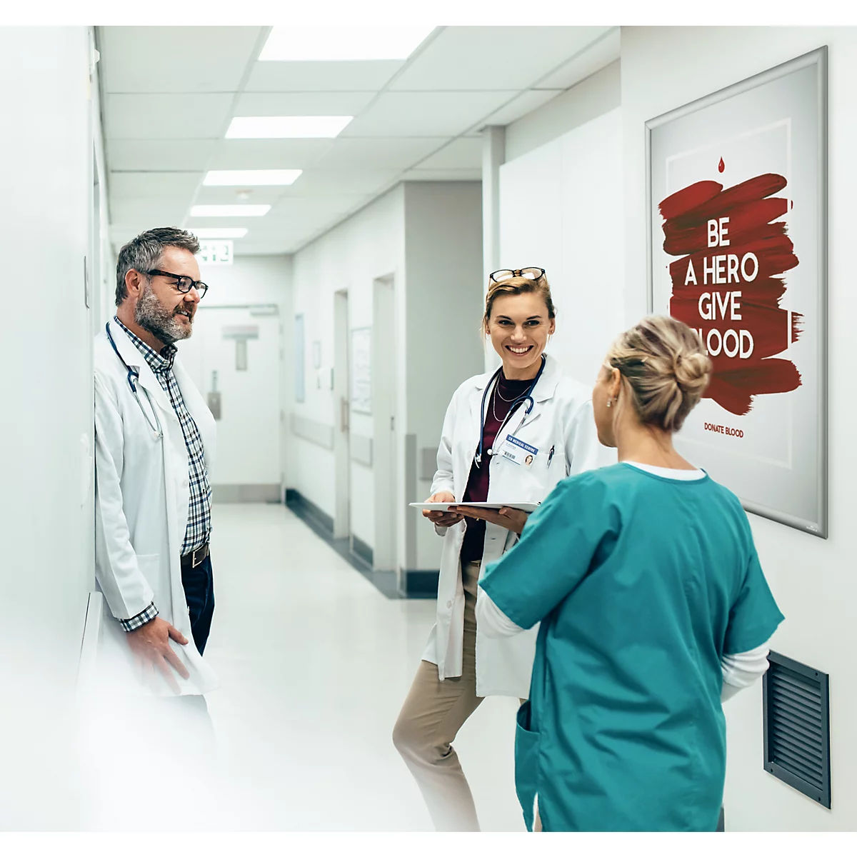 Drei medizinische Fachkräfte unterhalten sich freundlich auf einem hellen Krankenhausflur neben einem Blutspende-Plakat.