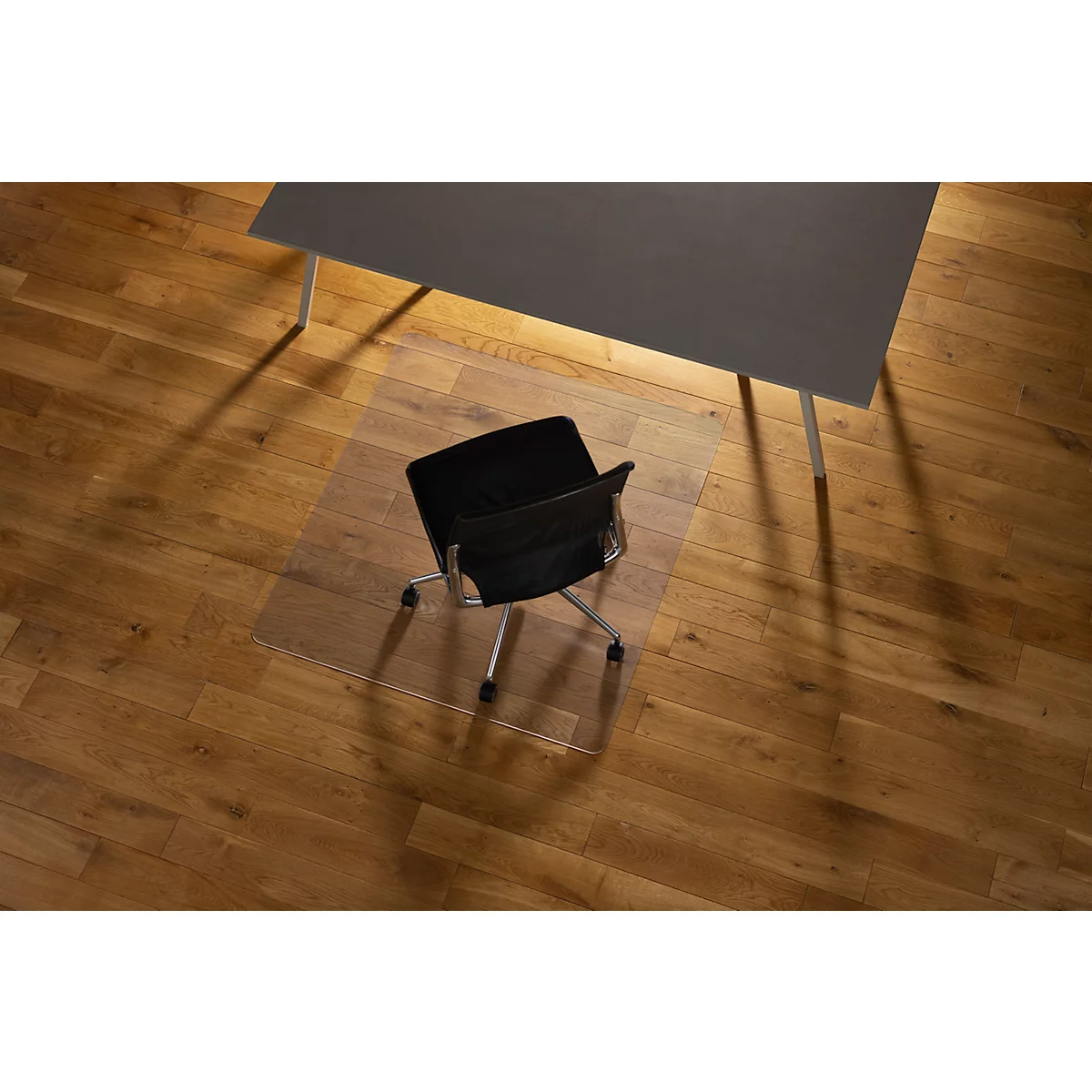 Silla de oficina sobre una alfombra de protección de suelo transparente debajo de un escritorio gris sobre un suelo de madera. Sombra.