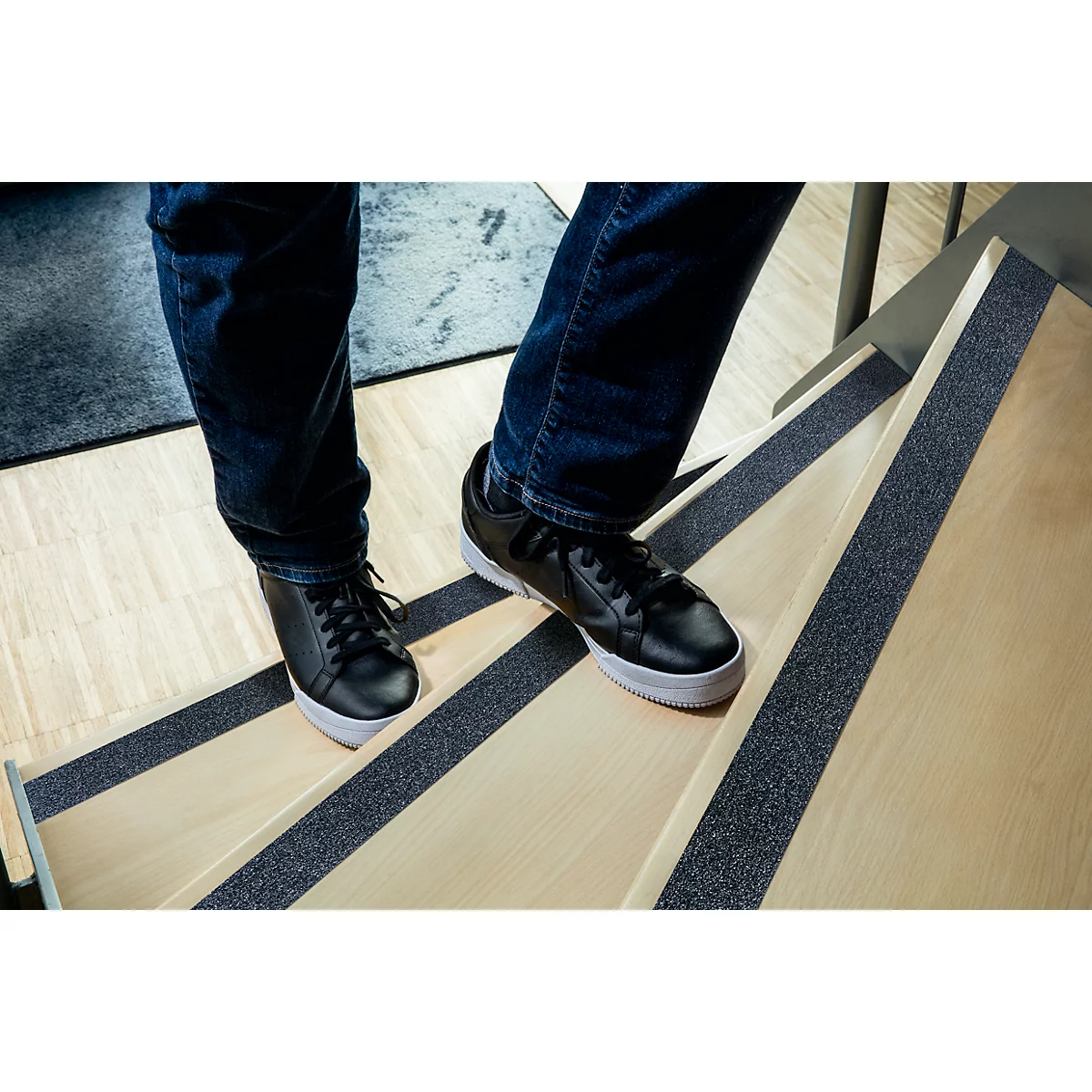 Person walking on wooden stairs. Black shoes, blue jeans. Black non-slip strips on steps.