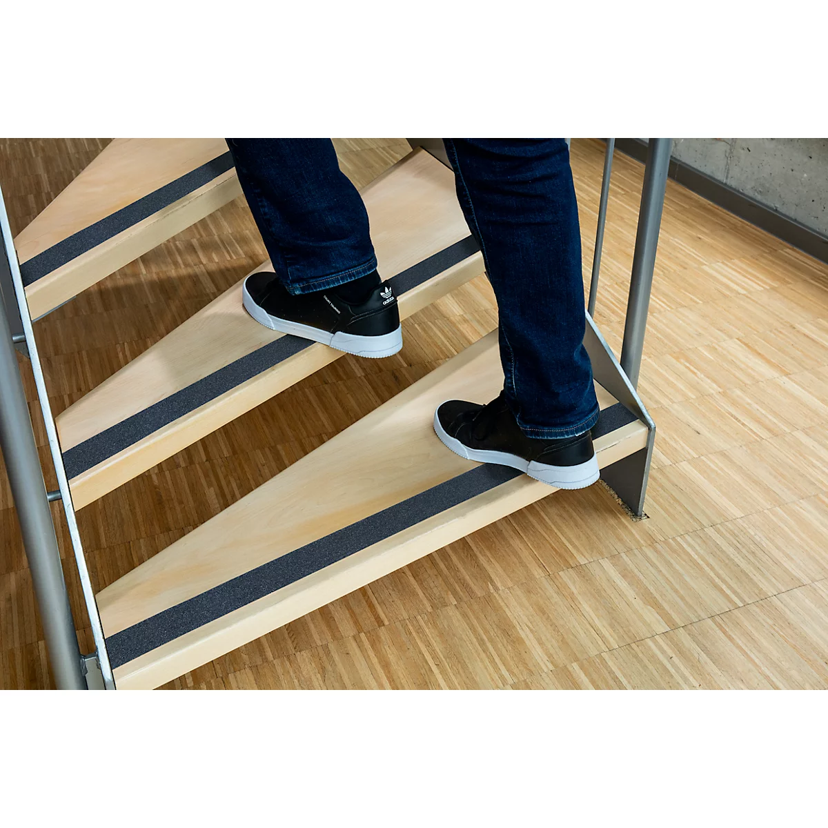 Person is walking up a wooden staircase with anti-slip strips. Black shoes with white soles. Blue jeans. Metal railing.