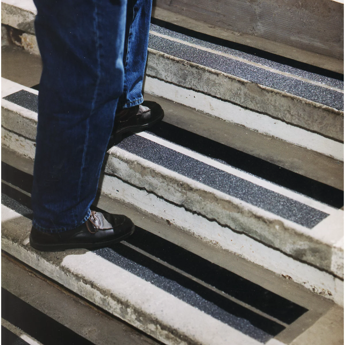 Person on stairs with non-slip strips. Black strips alternate with concrete. Jeans and black shoes.
