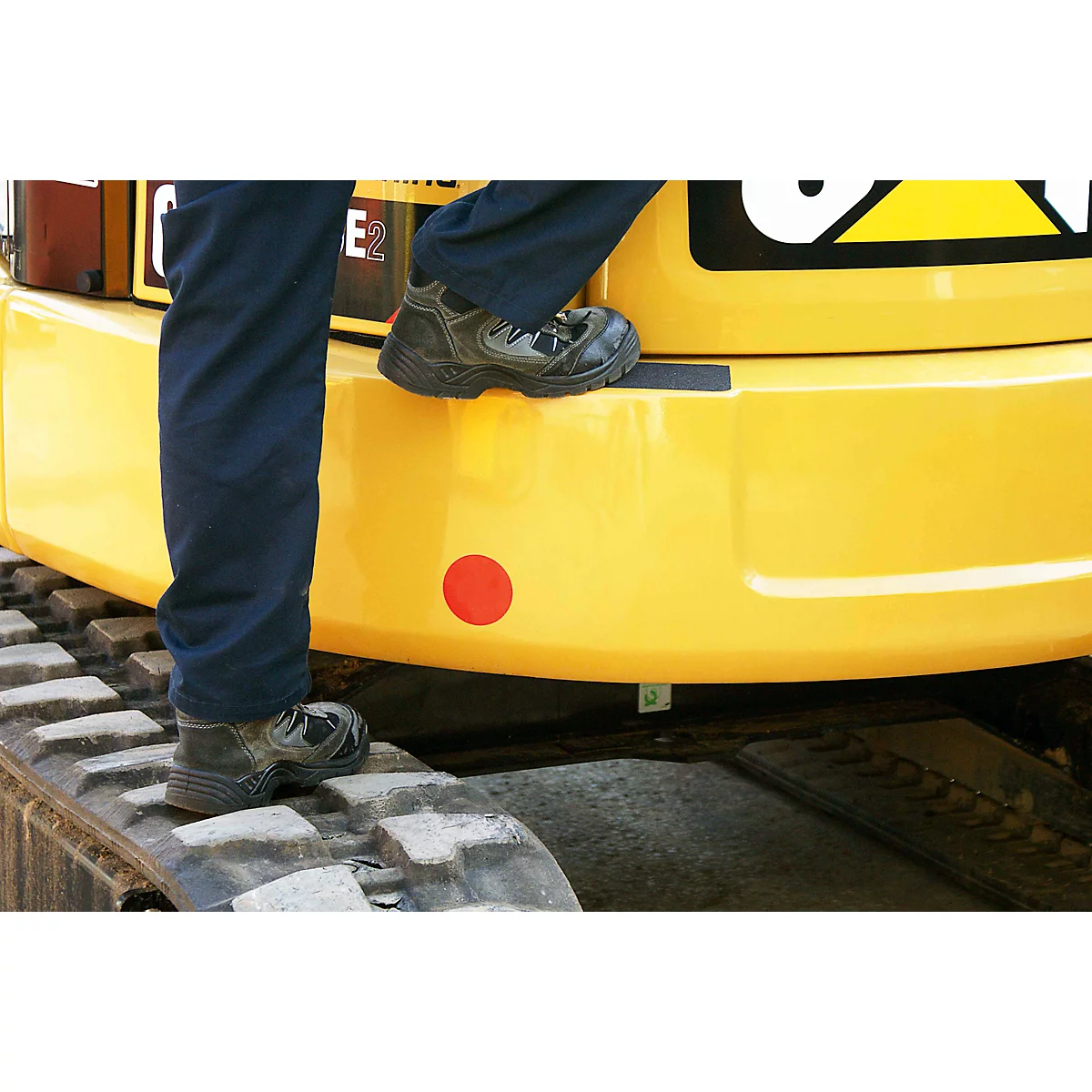 Close-up: worker stepping onto track of yellow excavator. Boots, legwear, detail. Red dot, black stripe.
