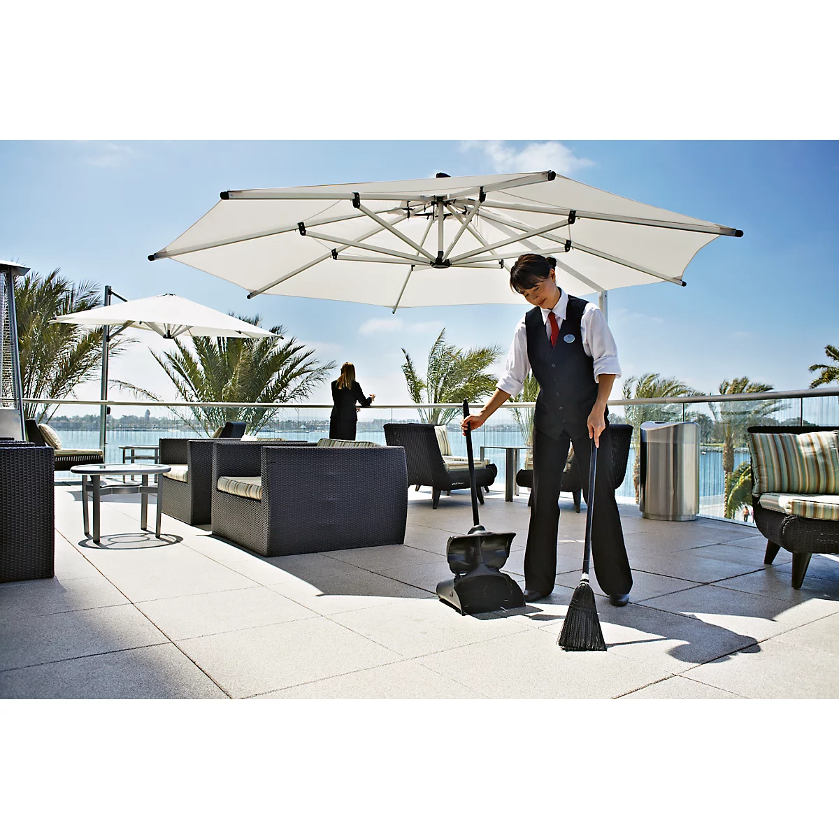 Waitress sweeping terrace under parasol, sea view. Another person stands in the background.