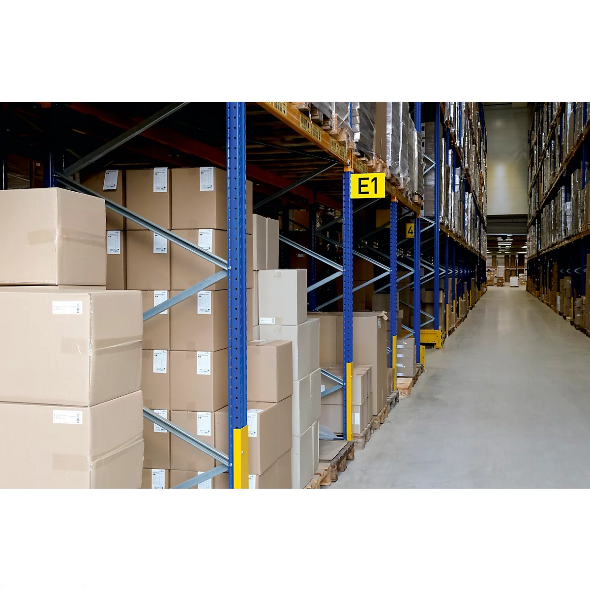 Rijen kartonnen dozen in een magazijnrek. Blauwe steunen, gele borden met letters en cijfers. Lichte betonnen vloer.