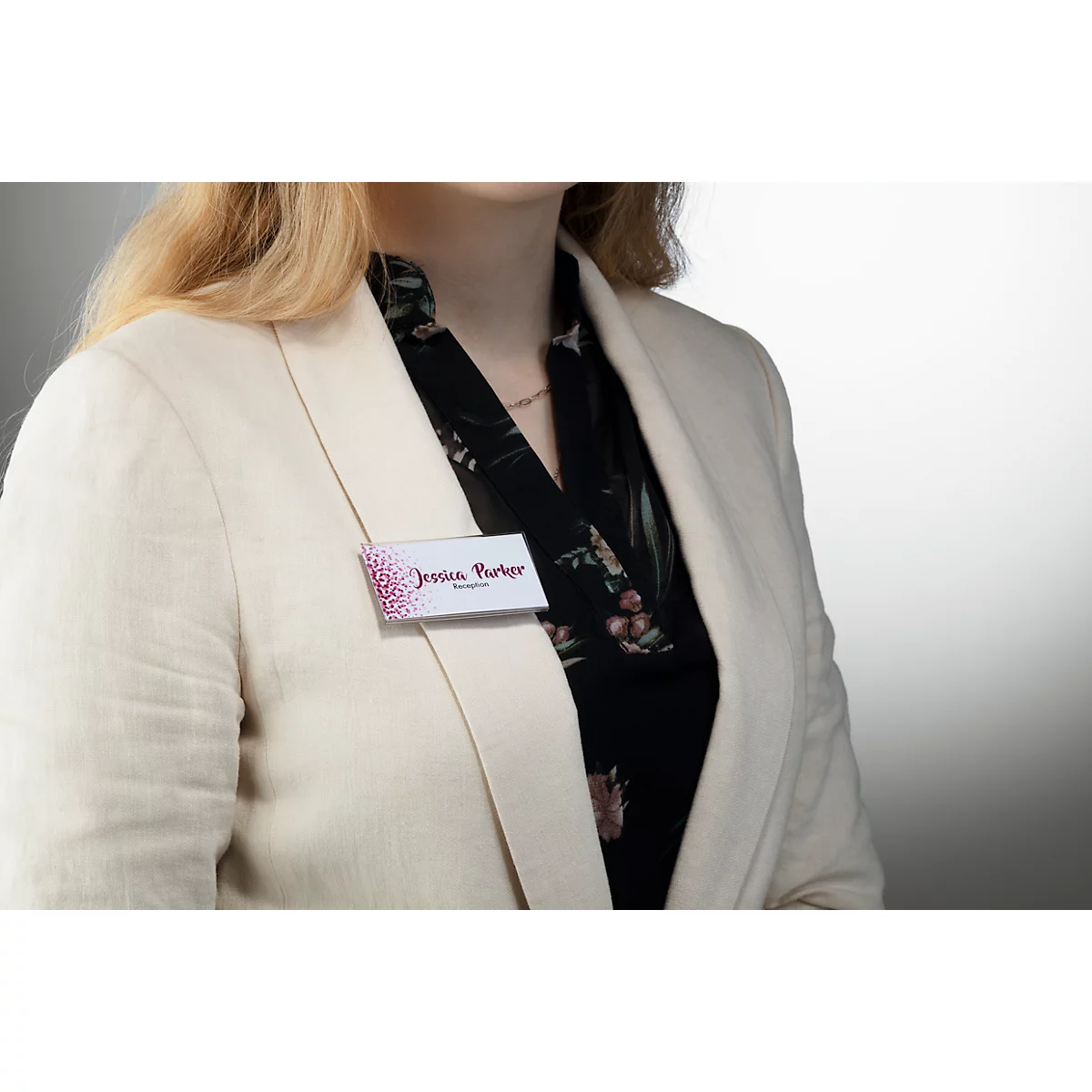 Persoon met naamkaart 'Jessica Parker' op blazer. Lichtblouse, zwart bloemenpatroon.