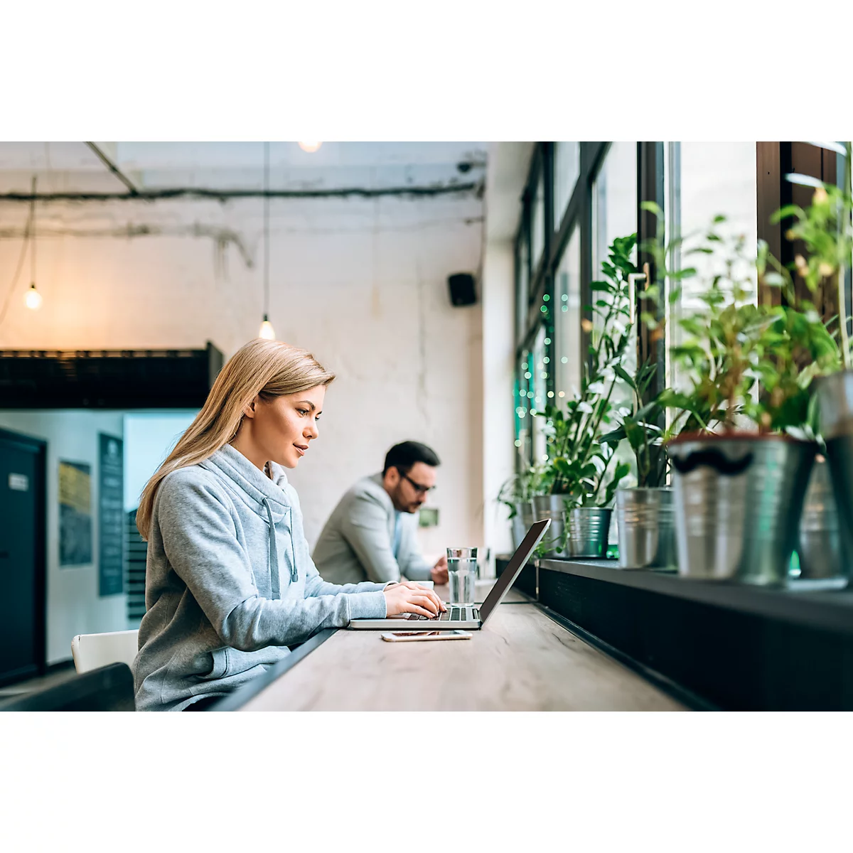 Vrouw typt op een laptop in een café. Op de achtergrond zit een man. Daarnaast potplanten bij het raam.