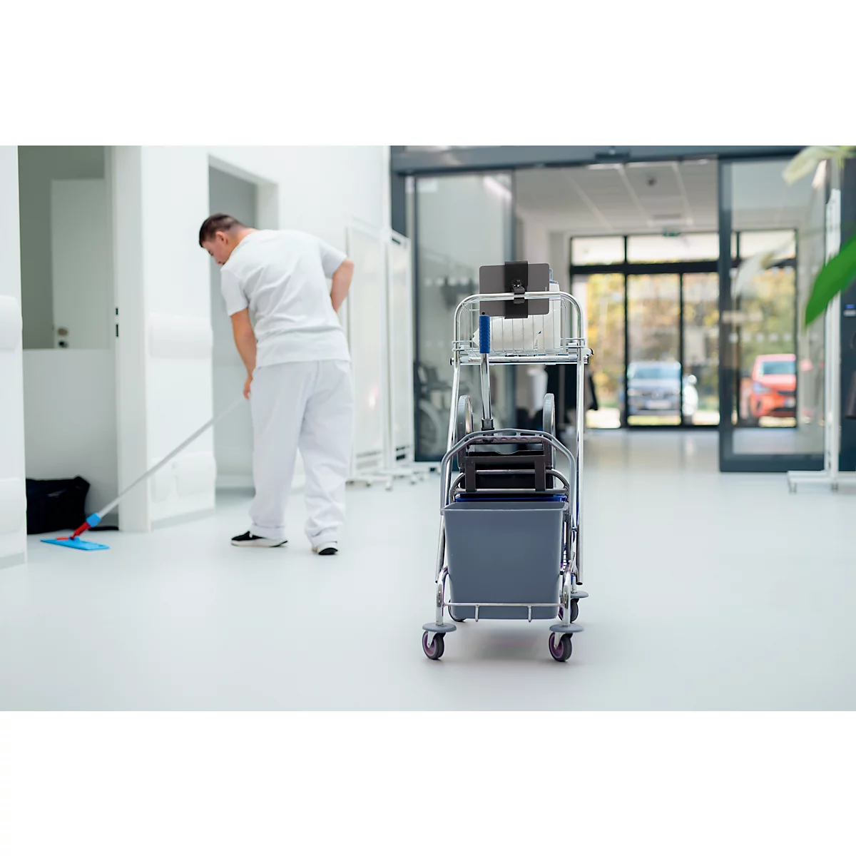 Een man maakt de vloer schoon in een helder gebouw. Een schoonmaakkar staat rechts.