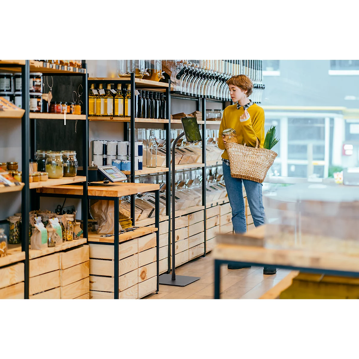 Jonge vrouw in een biologische winkel. Ze draagt een gele trui, jeans en een mandje. Schappen met voedsel.
